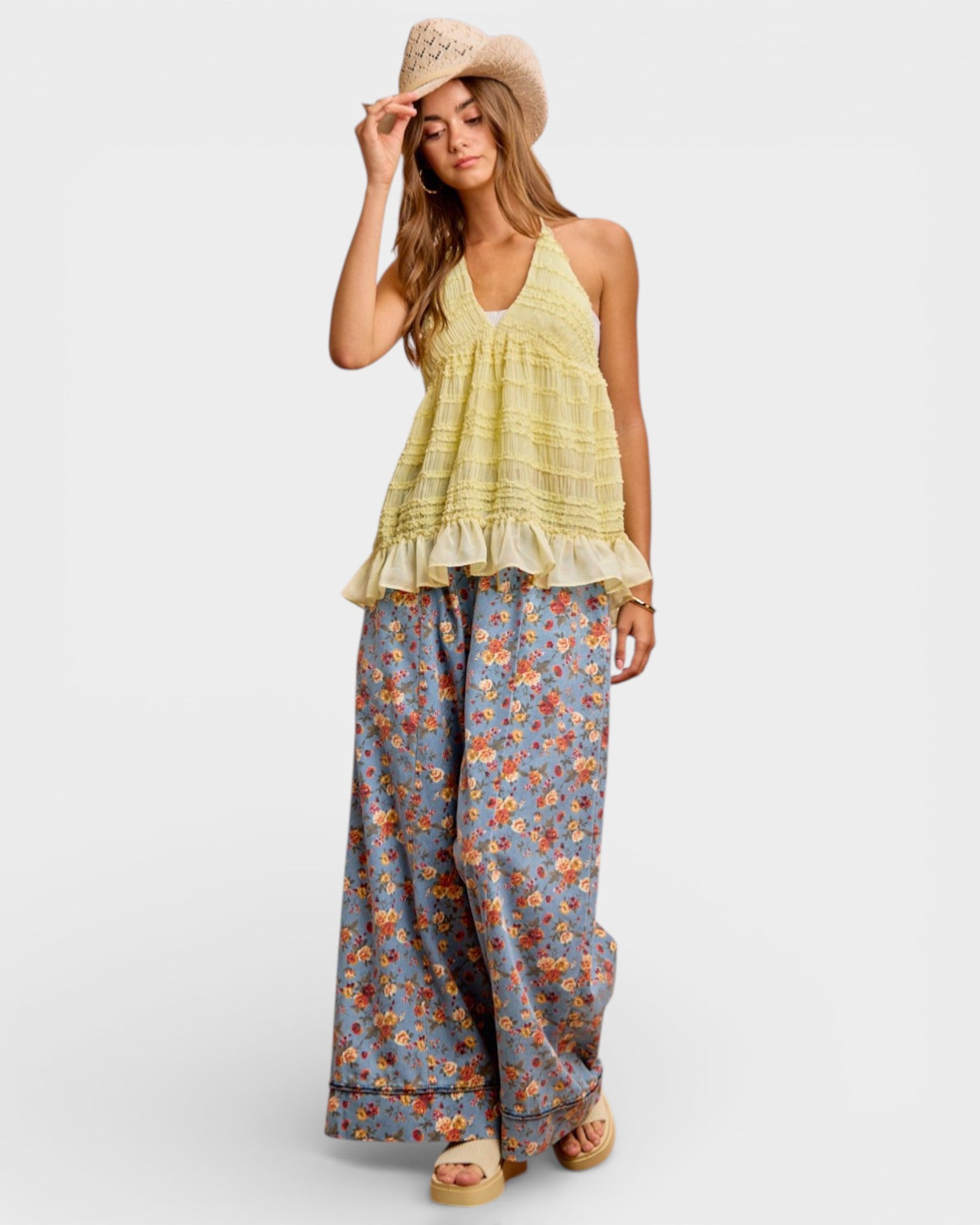 Woman wearing a yellow top and floral pants with a hat on a white background