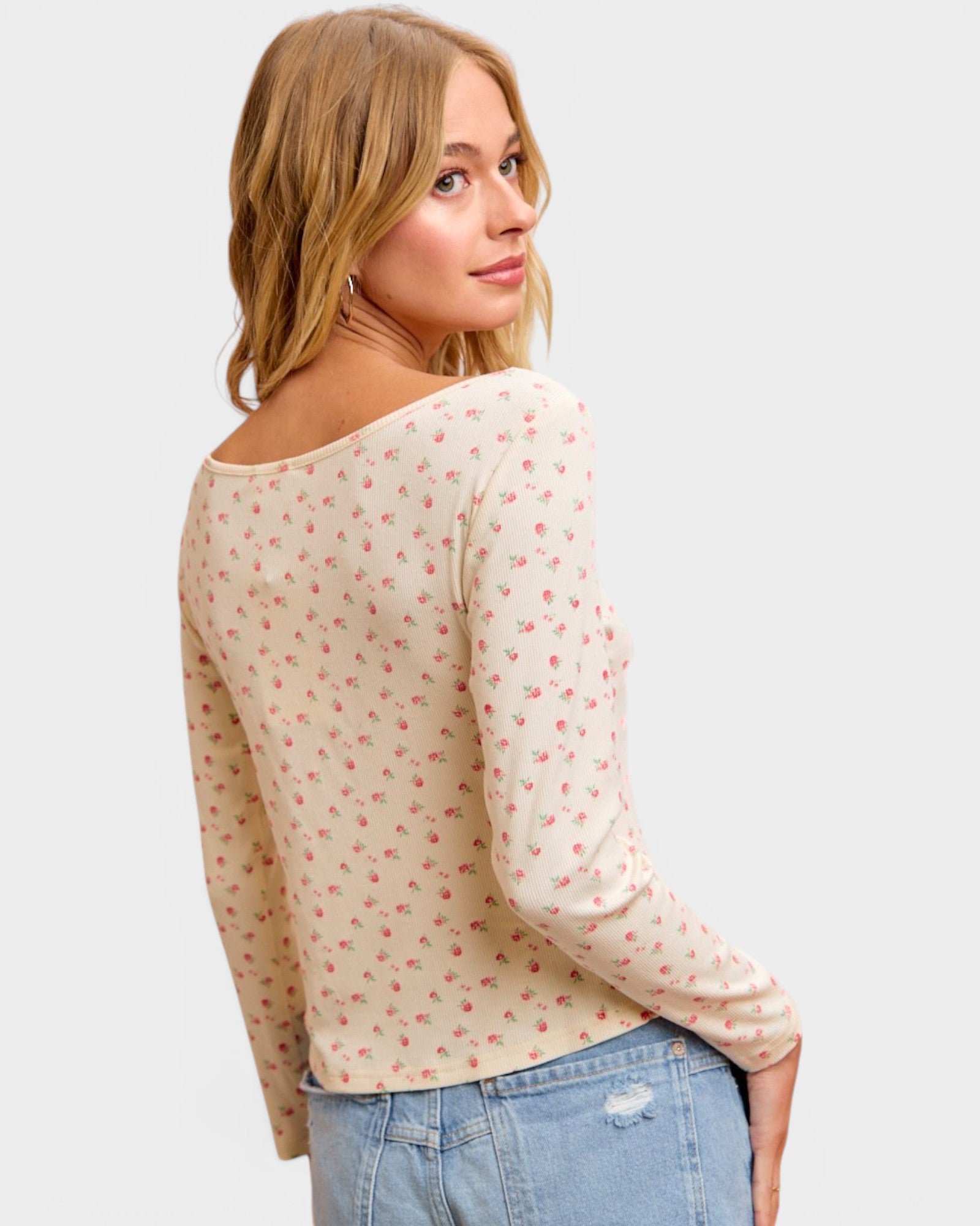 Woman wearing a cream long-sleeve top with red patterns and light blue jeans on a plain background