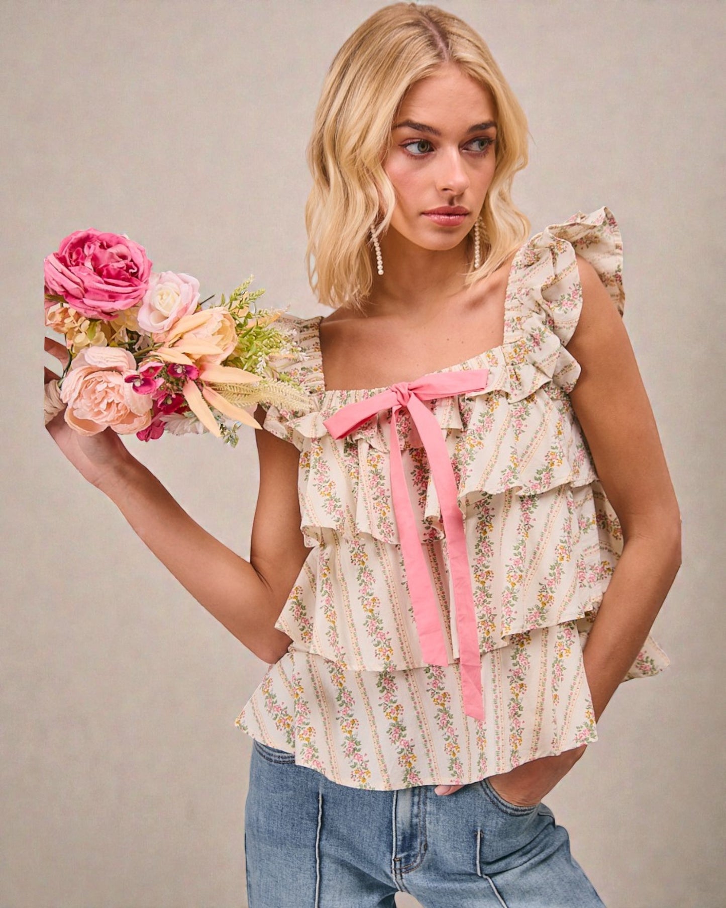 Woman wearing a floral blouse with ruffles and a pink ribbon, holding flowers against a neutral background