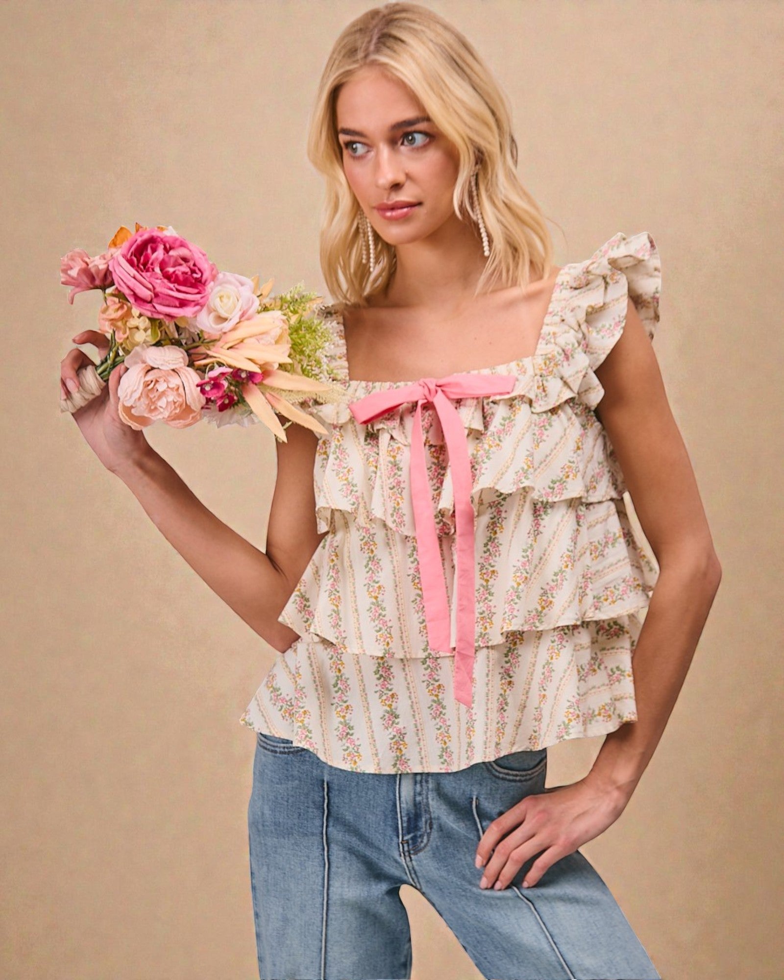 Woman wearing a floral top with ruffles and a pink ribbon, holding flowers against a beige background