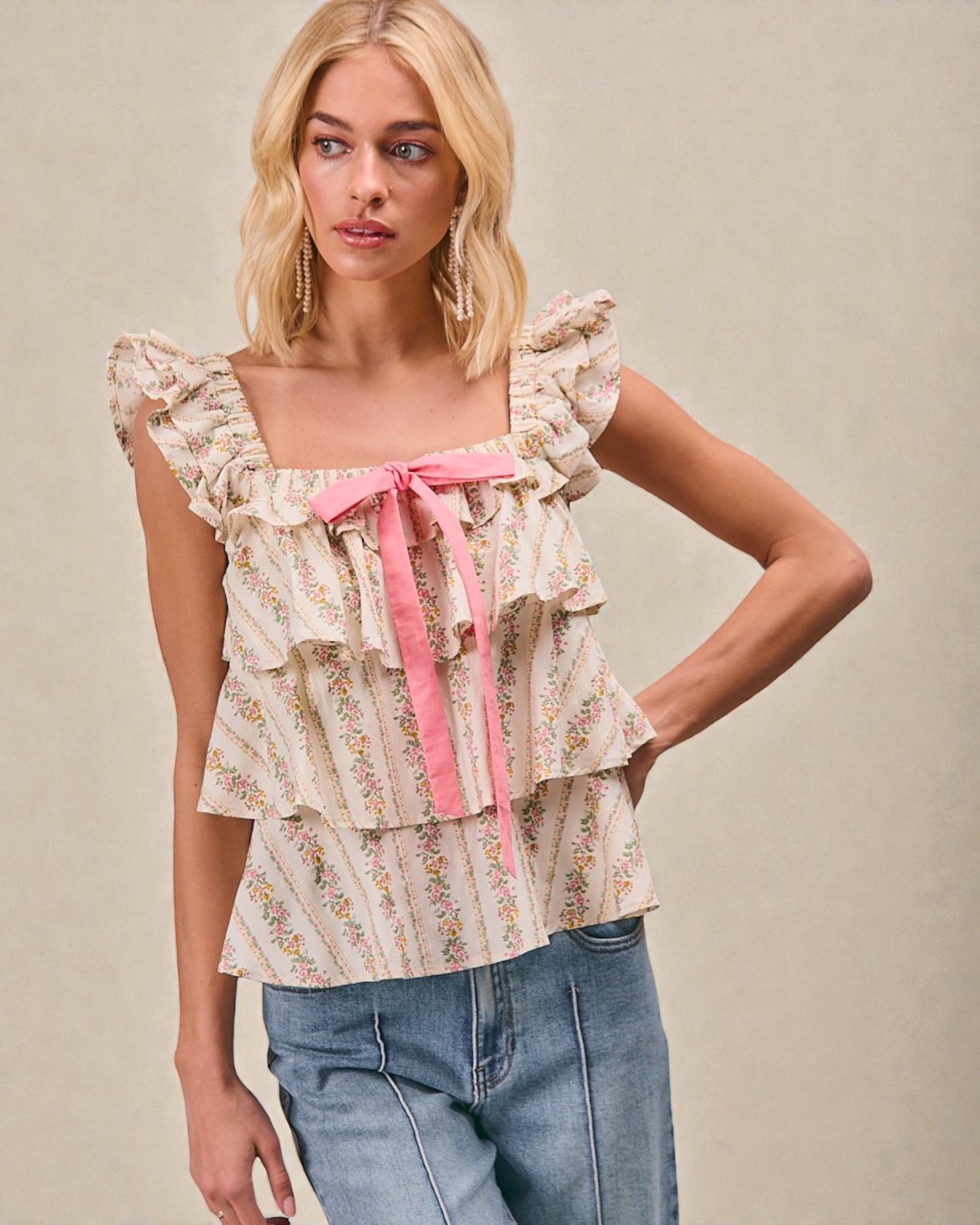 Woman wearing a floral blouse with ruffles and a pink ribbon, standing against a beige background.