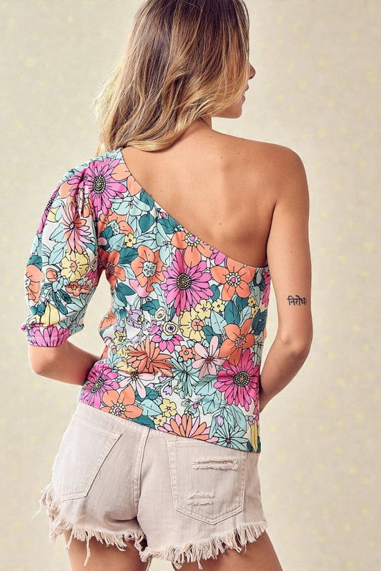 Woman wearing a colorful floral one-shoulder top and light shorts against a neutral background