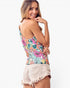 A woman wearing a colorful floral print tank top and light beige frayed denim shorts, photographed against a plain white studio background.