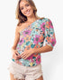 A woman wearing a one-shoulder short-sleeve top with a colorful floral print, paired with light beige denim shorts, against a plain white background.