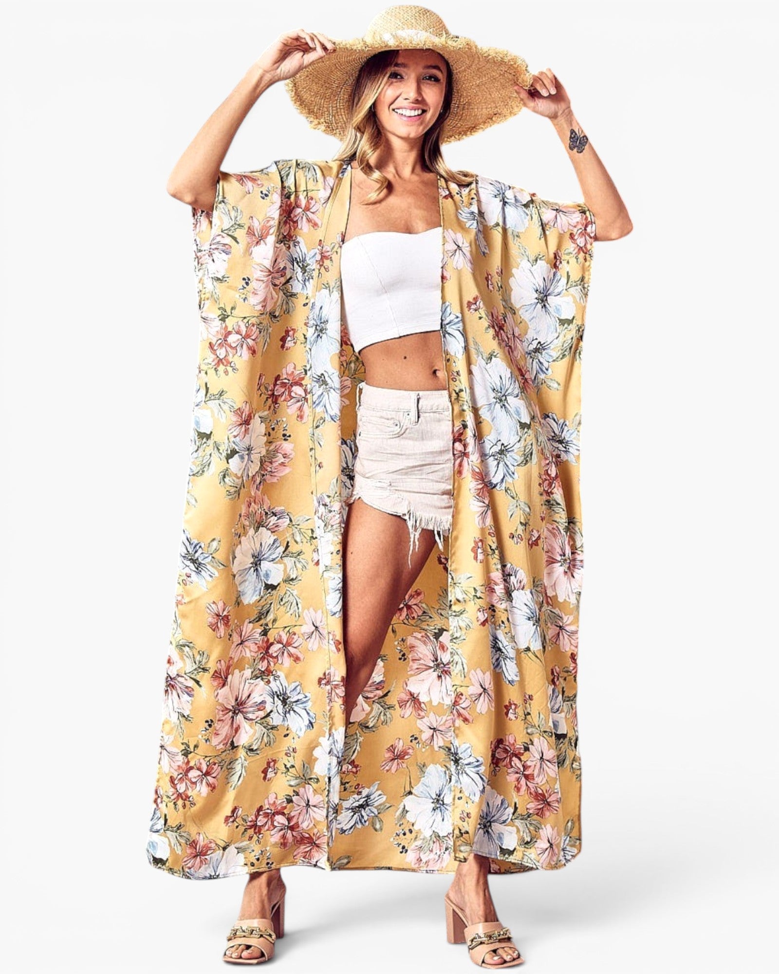 A woman modeling a long, open-front kimono-style cover-up with a yellow base and a multicolor floral print, worn over a white crop top and light denim shorts, with a wide-brim straw hat and sandals.