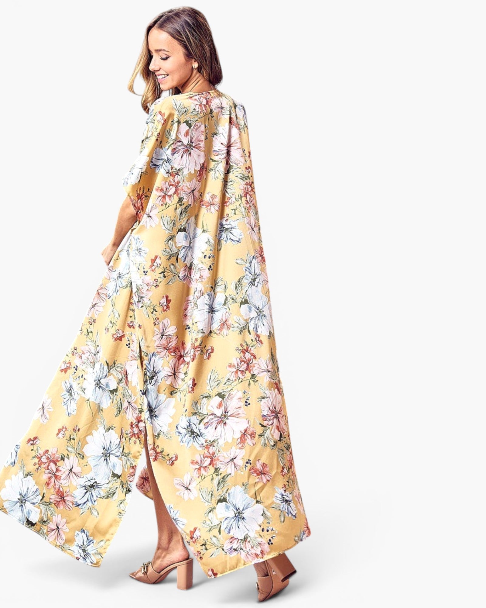 A woman modeling a long, flowing yellow floral dress or caftan with a side slit, shown from a three-quarter back angle against a plain light gray studio background.