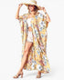 A woman modeling a long, flowing yellow floral kimono cover-up with a white strapless top, white denim shorts, and a straw hat against a light gray studio background.