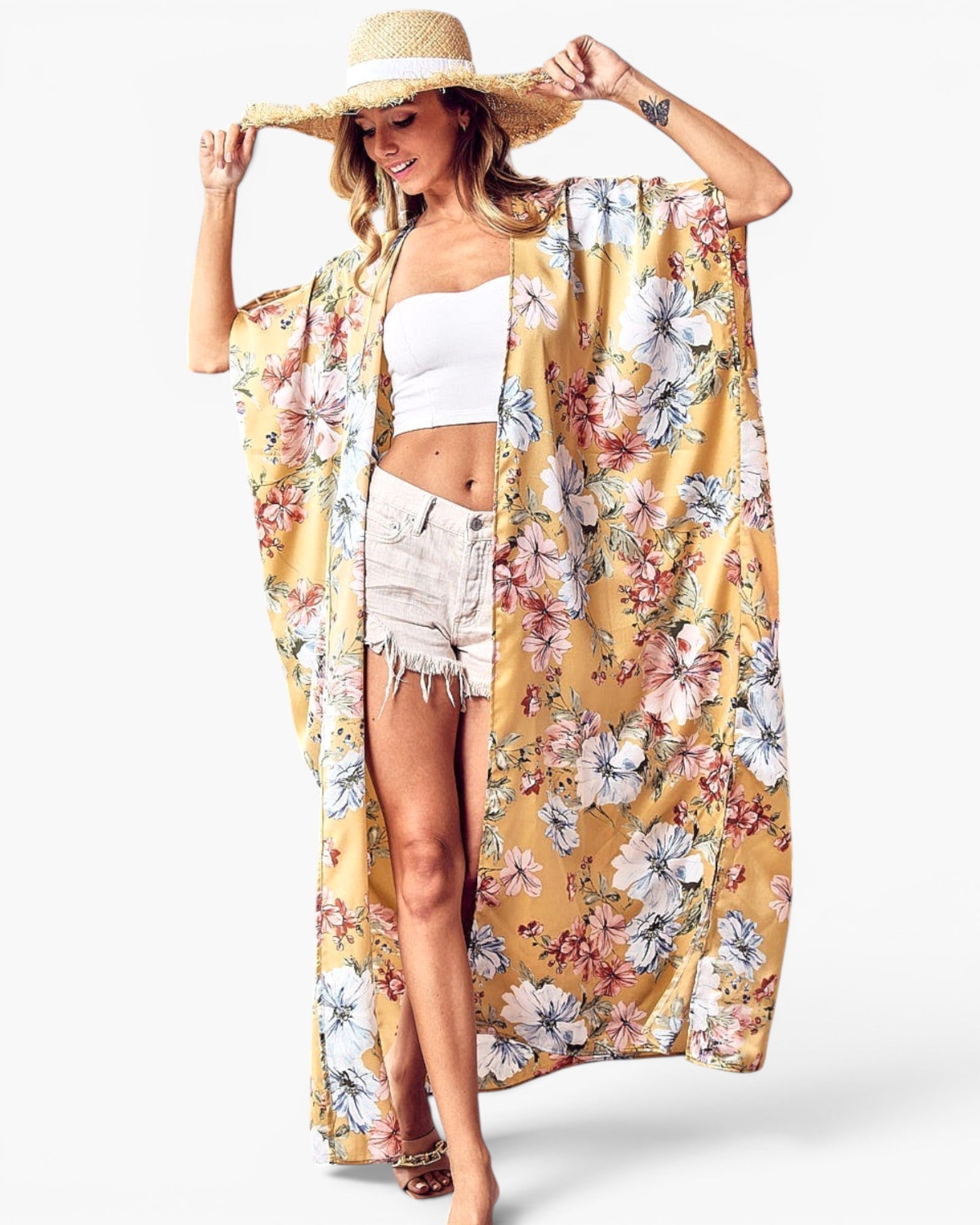 A woman modeling a long, open-front floral kimono cover-up in mustard yellow with large white, pink, and green flowers, paired with a straw hat, white crop top, and denim shorts against a light gray studio background.