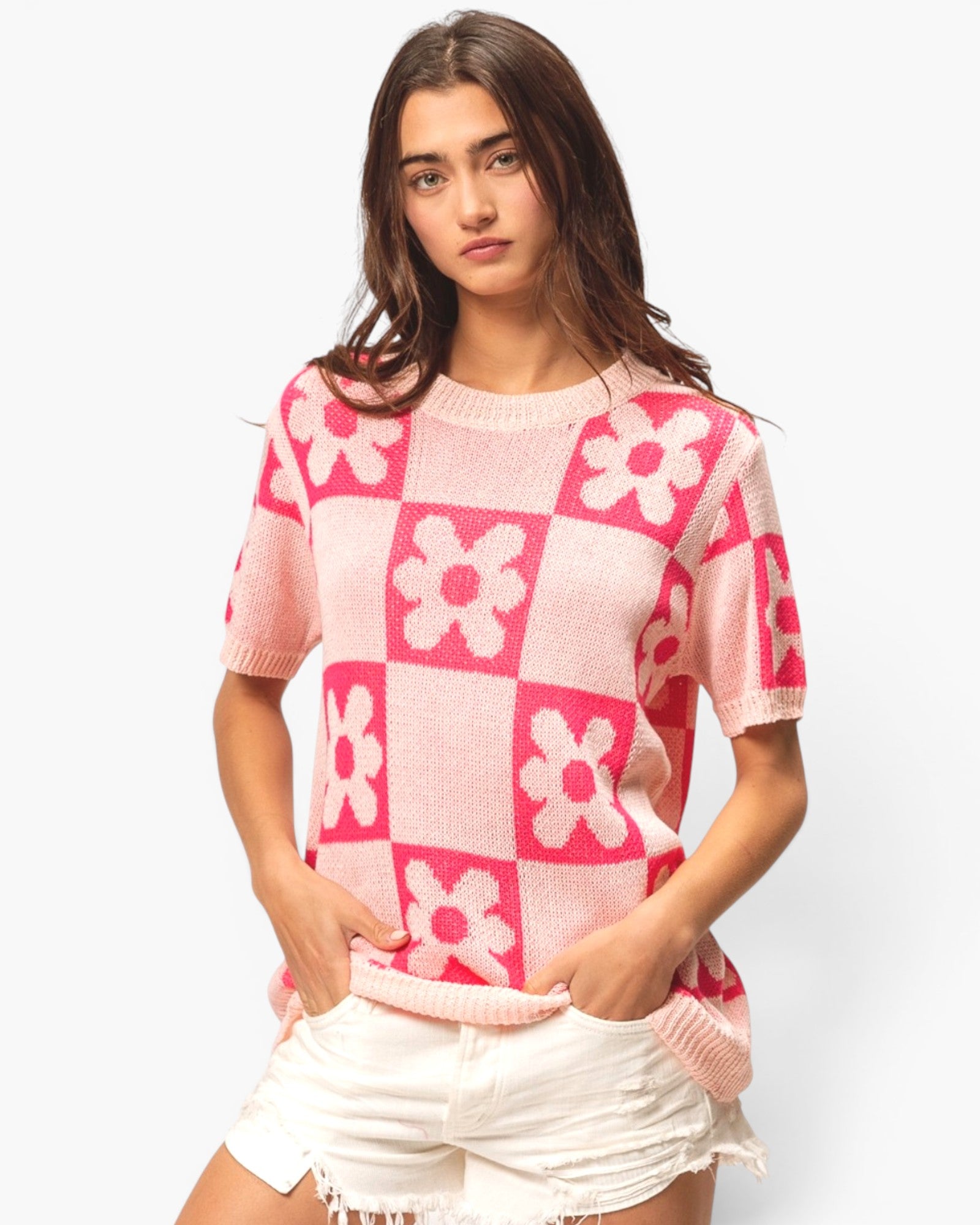 A woman modeling a pink and white knit short-sleeve sweater with a checkerboard and flower pattern, paired with white shorts, against a light gray studio background.