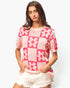 A woman modeling a pink and white knit short-sleeve sweater with a checkerboard and flower pattern, paired with white shorts, against a light gray studio background.