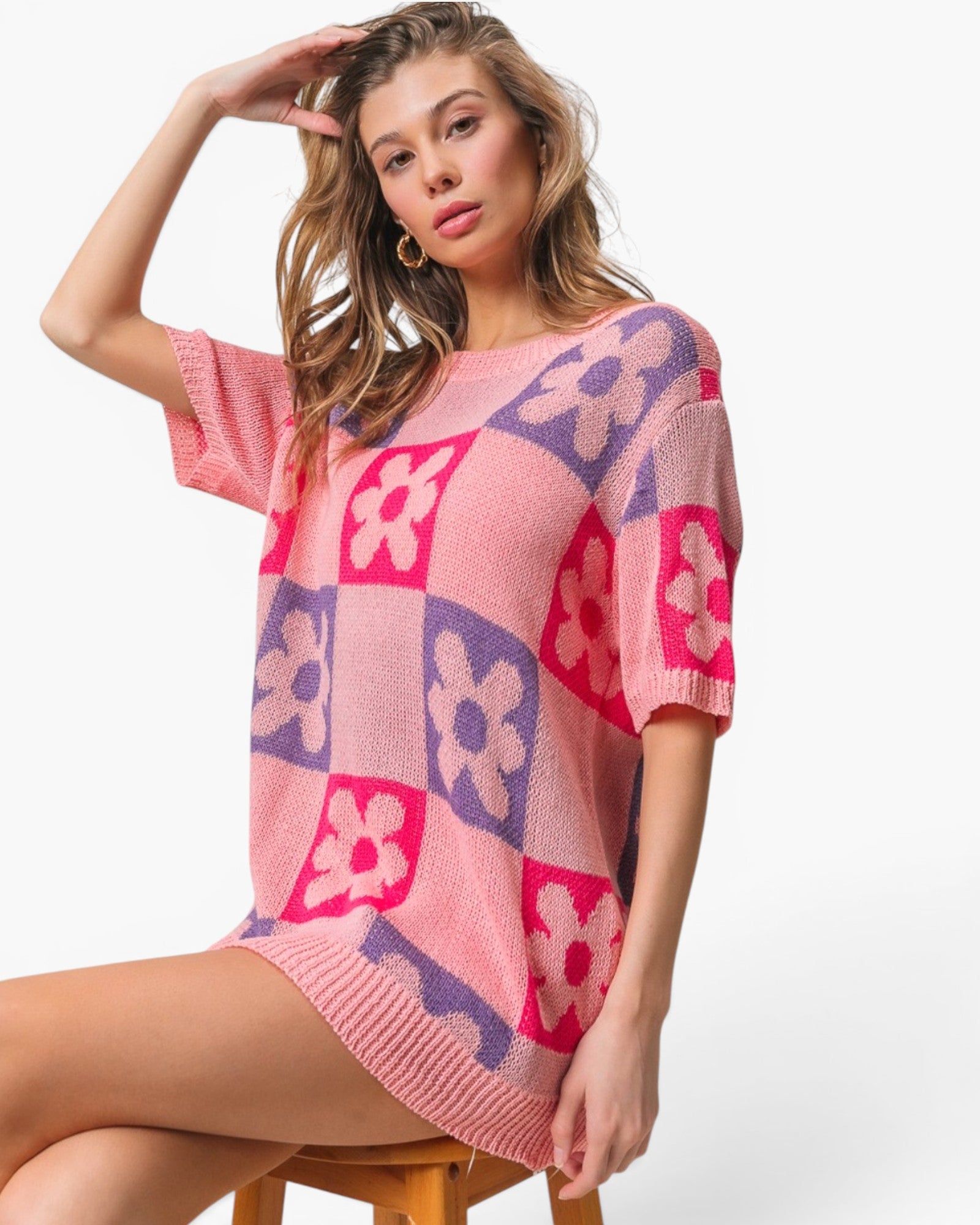 A woman models a pink knit mini dress with a retro checkerboard pattern of pink and purple squares and floral motifs, sitting on a wooden stool against a white studio background.