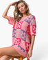 A woman models a pink knit mini dress with a retro checkerboard pattern of pink and purple squares and floral motifs, sitting on a wooden stool against a white studio background.