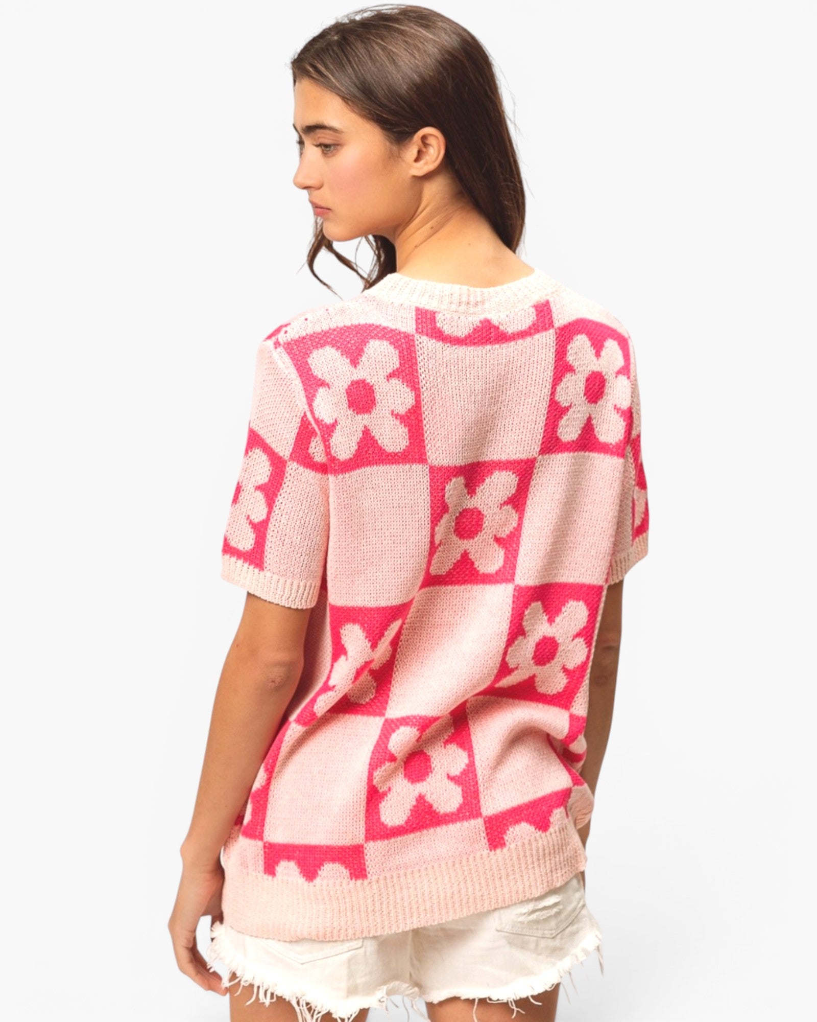 A woman shown from the back wearing a pink and cream knit sweater with a checkerboard pattern and raised flower motifs, paired with white frayed shorts against a light gray studio background.