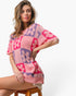 A woman wearing a pink and purple knit short-sleeve sweater with a floral patchwork pattern, seated on a wooden stool against a plain white background.