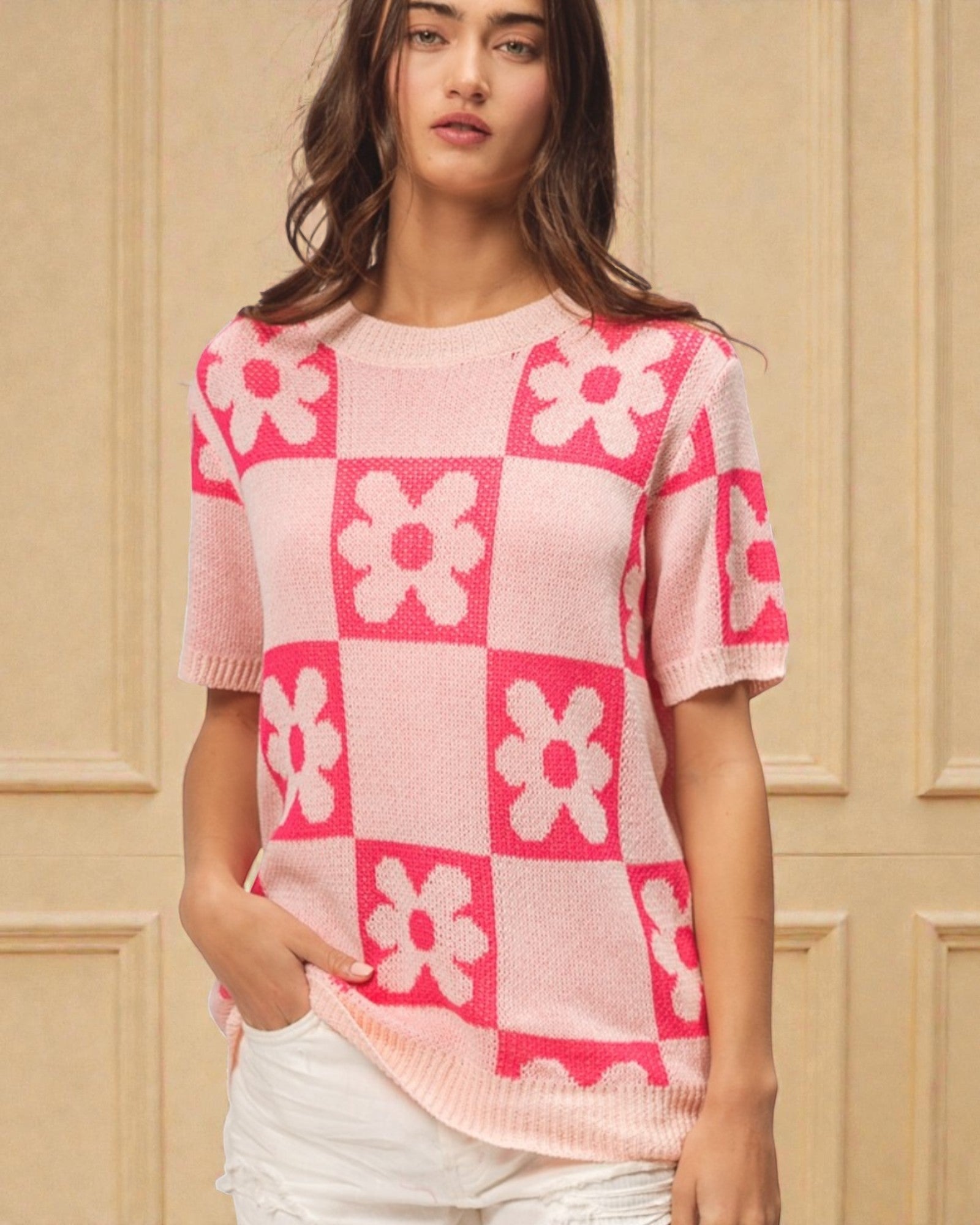 Woman wearing a pink and white checkered sweater with floral patterns against a beige wall.