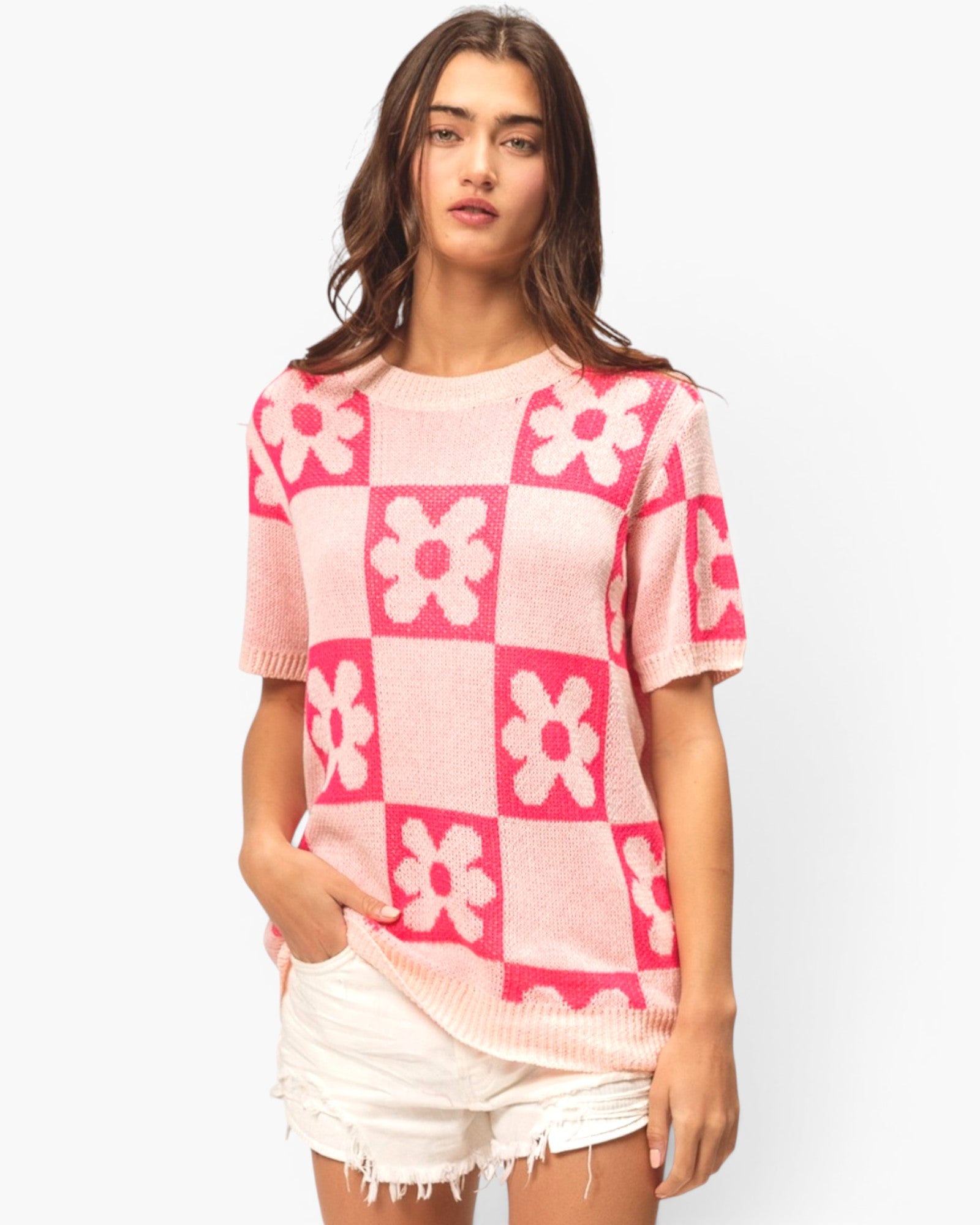 A woman wearing a pink and white checkerboard knit sweater with a floral pattern, paired with white frayed denim shorts, against a light gray studio background.