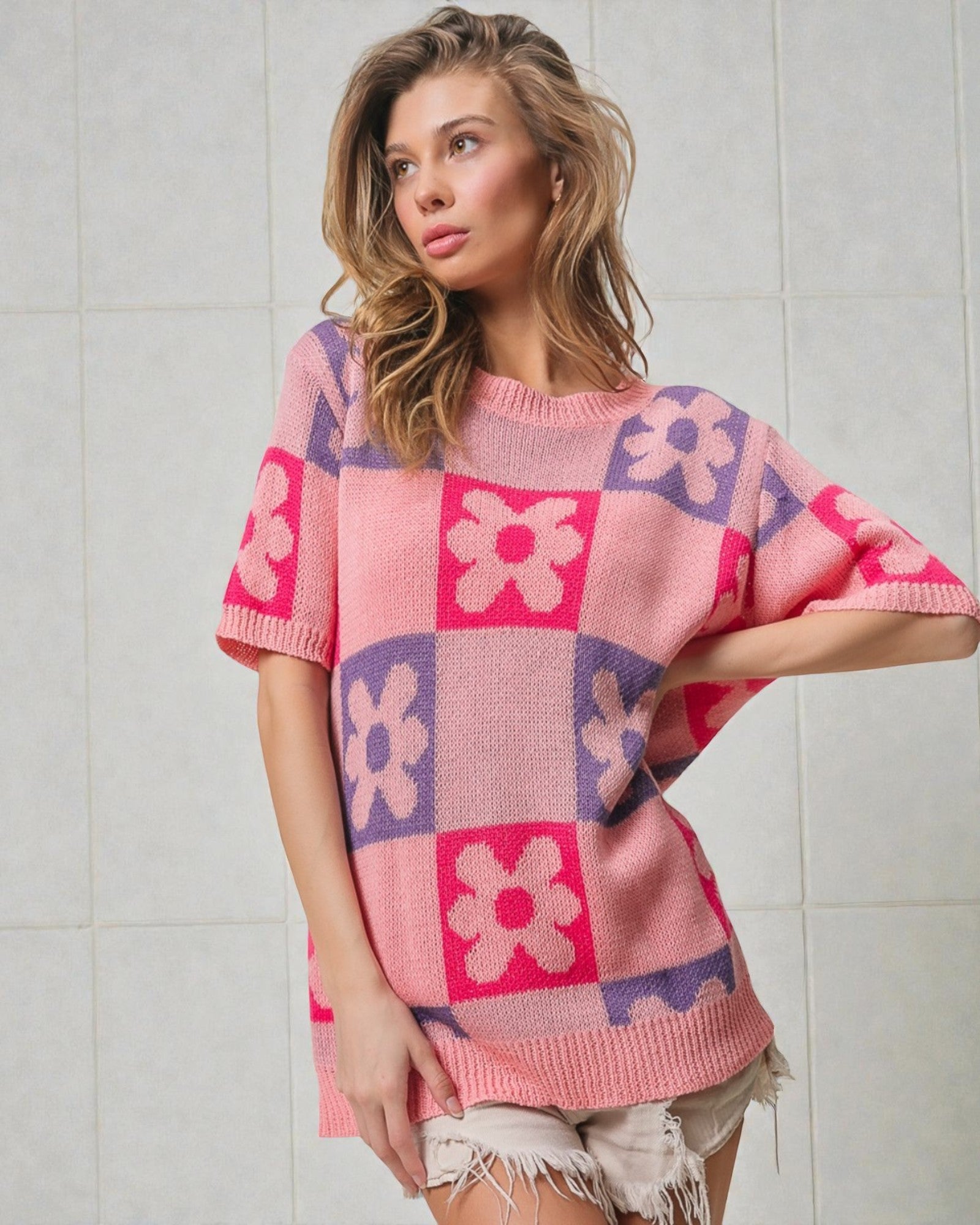 Woman wearing a pink and purple checkered sweater with floral patterns against a tiled wall.