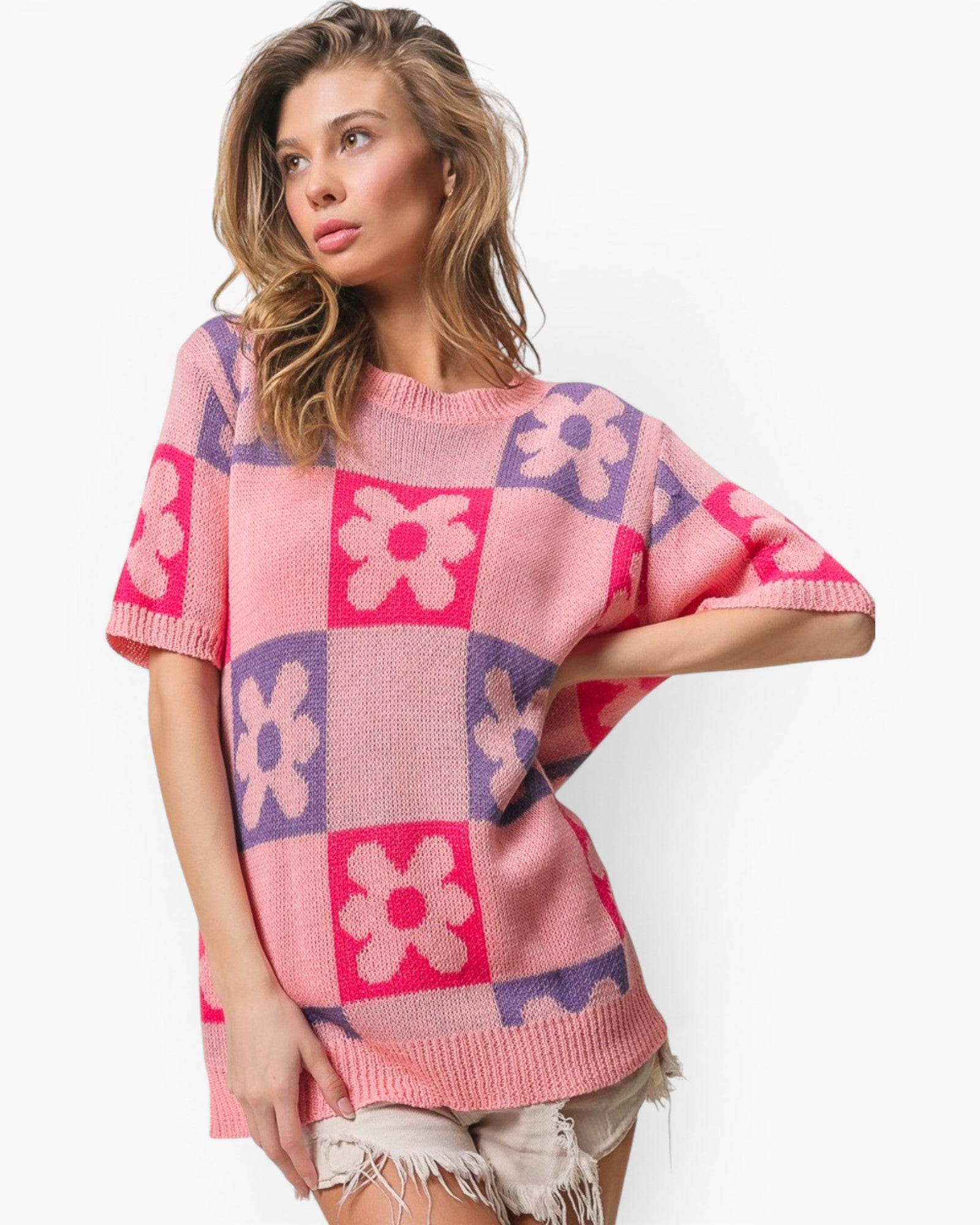 A woman wearing a pink knit sweater with a purple and pink checkerboard pattern and large flower motifs, paired with distressed light beige shorts, against a plain white background.