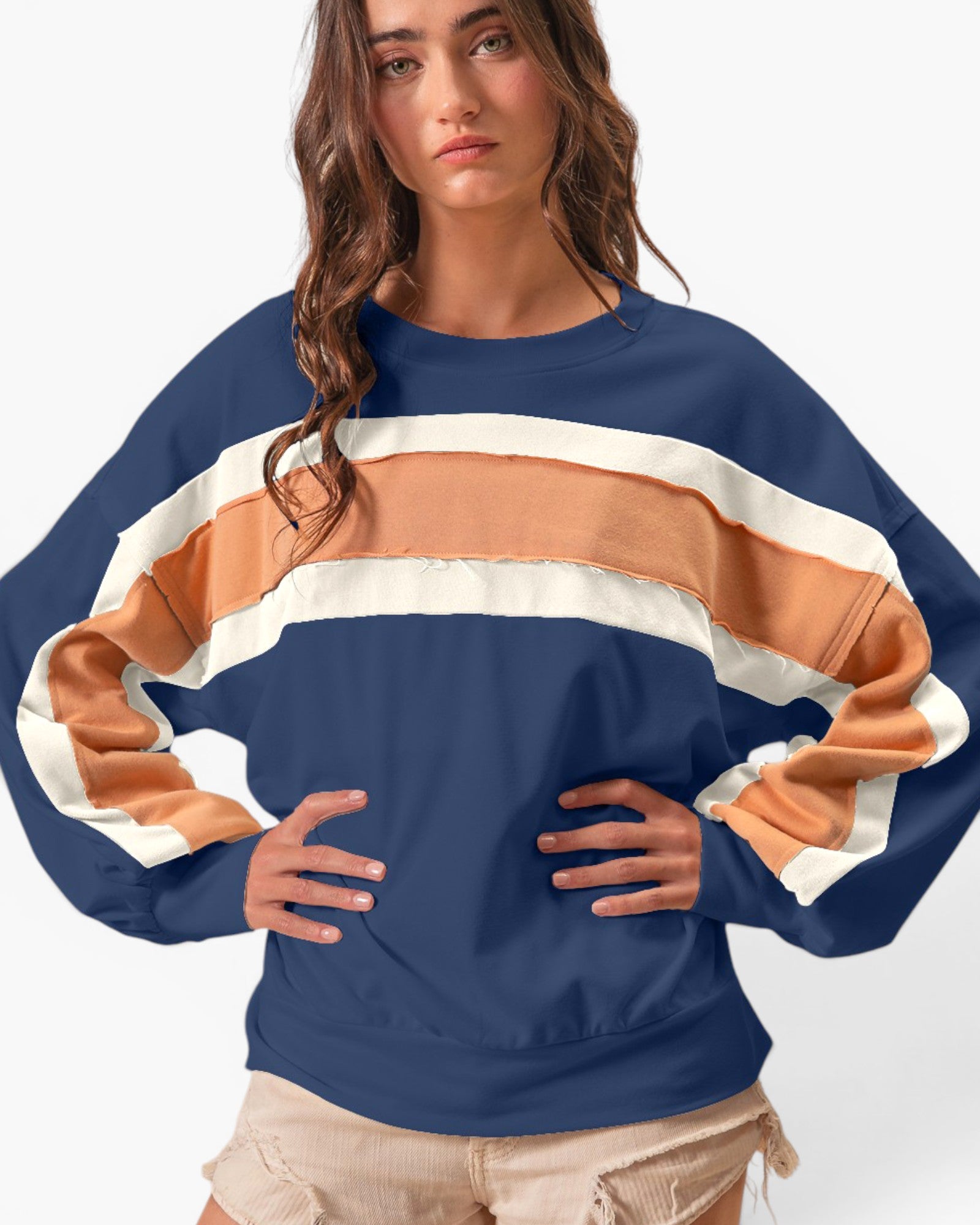 A woman wearing a navy blue sweatshirt with wide, draped sleeves and horizontal stripes in tan and off-white across the chest and sleeves, paired with beige shorts against a plain white background.