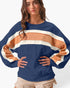 A woman wearing a navy blue sweatshirt with wide, draped sleeves and horizontal stripes in tan and off-white across the chest and sleeves, paired with beige shorts against a plain white background.