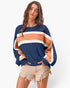 A woman modeling a loose long-sleeve sweatshirt with horizontal color-blocked stripes in navy blue, peach, and cream, paired with frayed beige denim shorts against a light gray studio background.