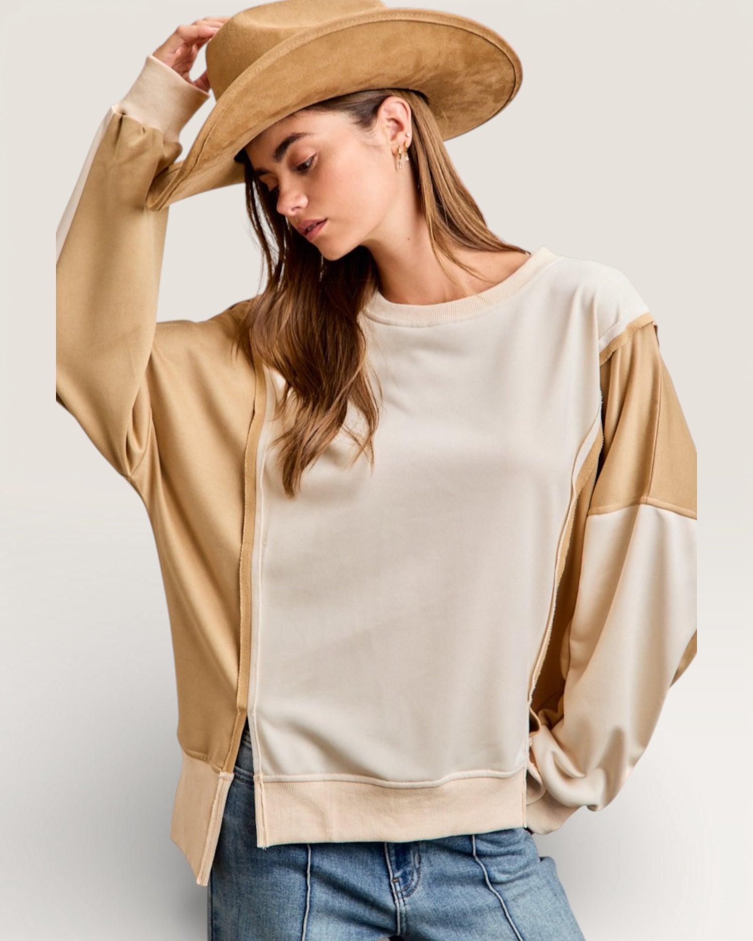 Woman wearing a beige and brown color-blocked sweater with a wide-brimmed hat on a plain background