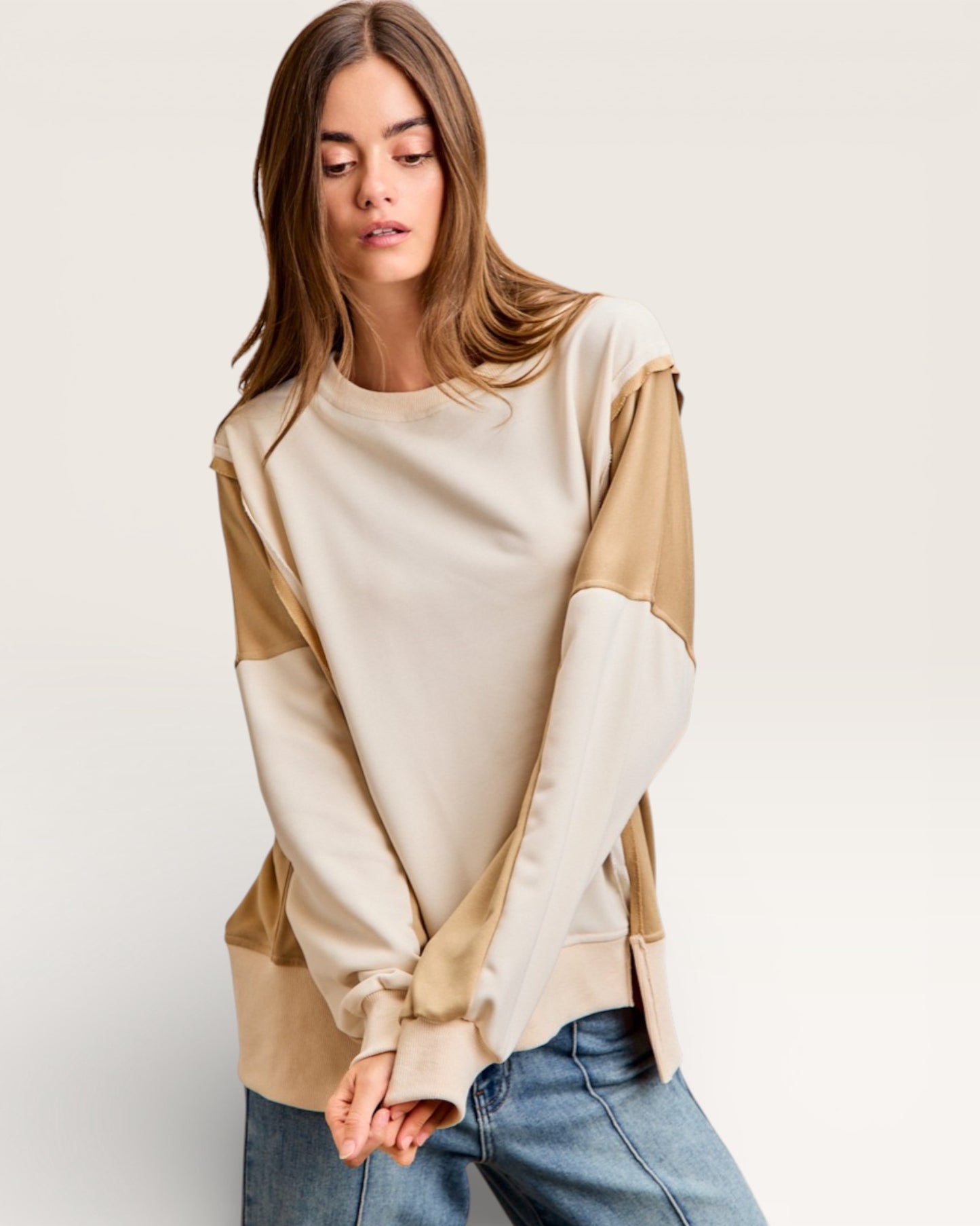 Woman wearing a beige and white color-blocked sweater with blue jeans on a plain background