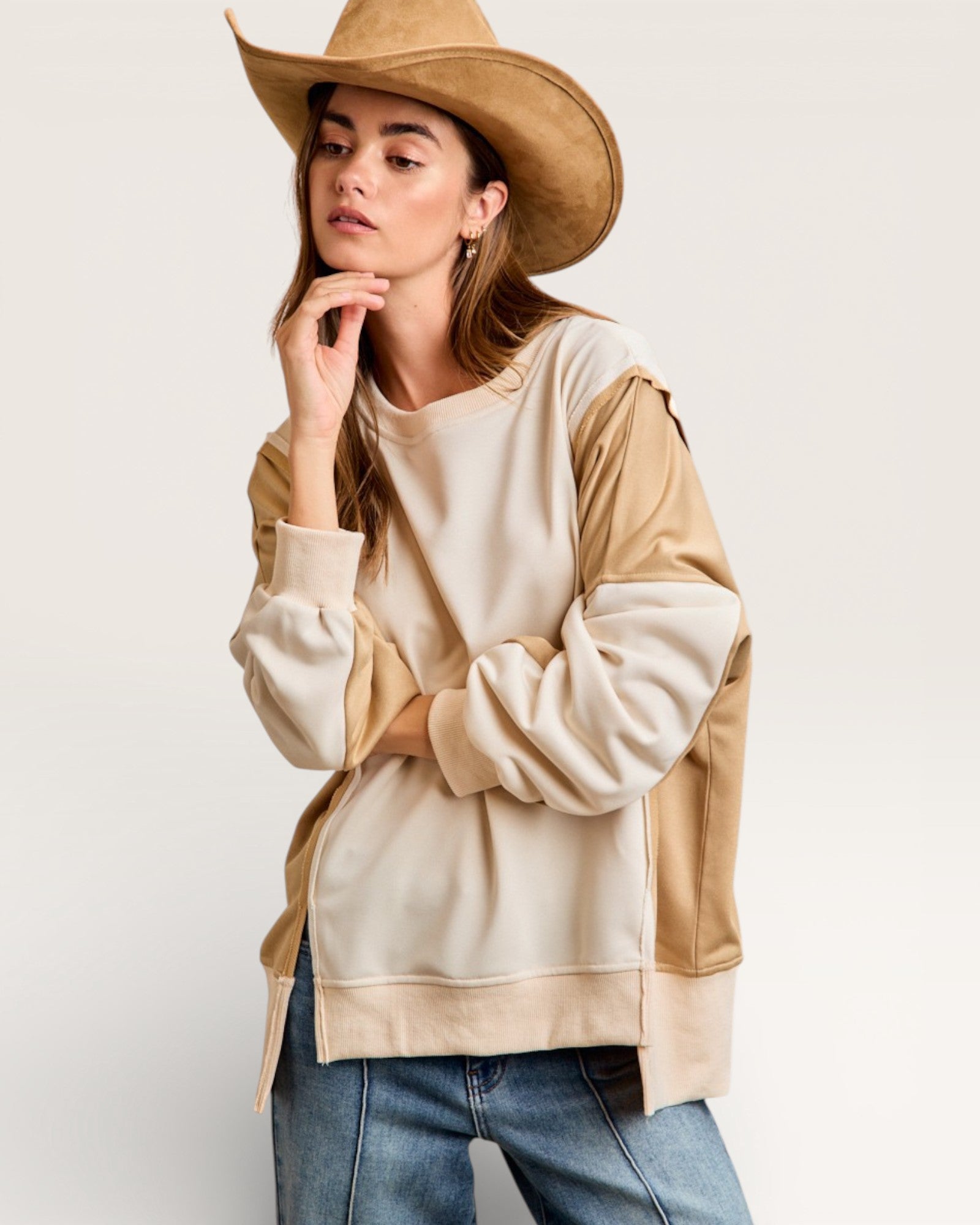 Woman wearing a beige and brown color-blocked sweatshirt with a wide-brimmed hat on a plain background