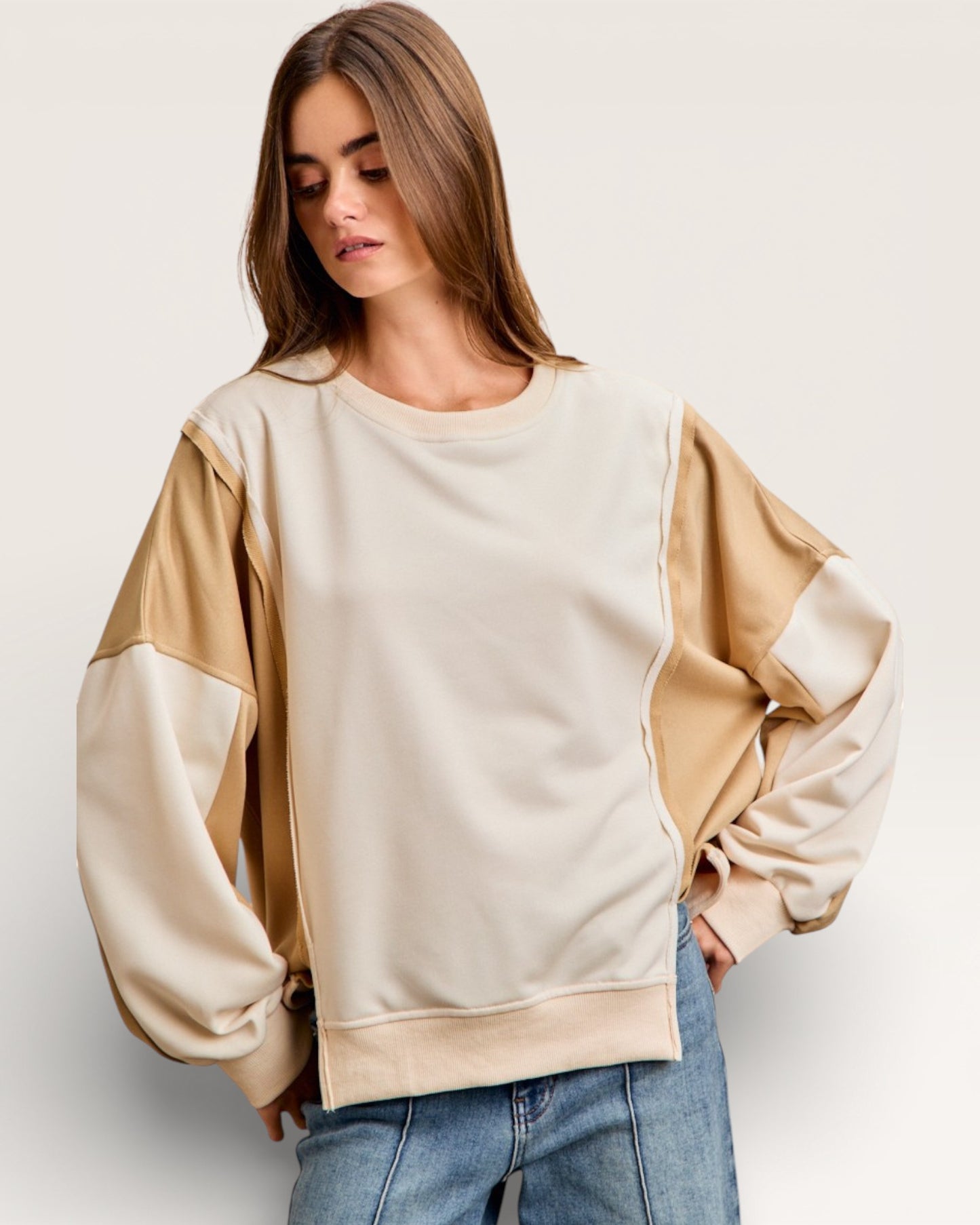 Woman wearing a beige and brown color-blocked sweater with blue jeans on a plain background