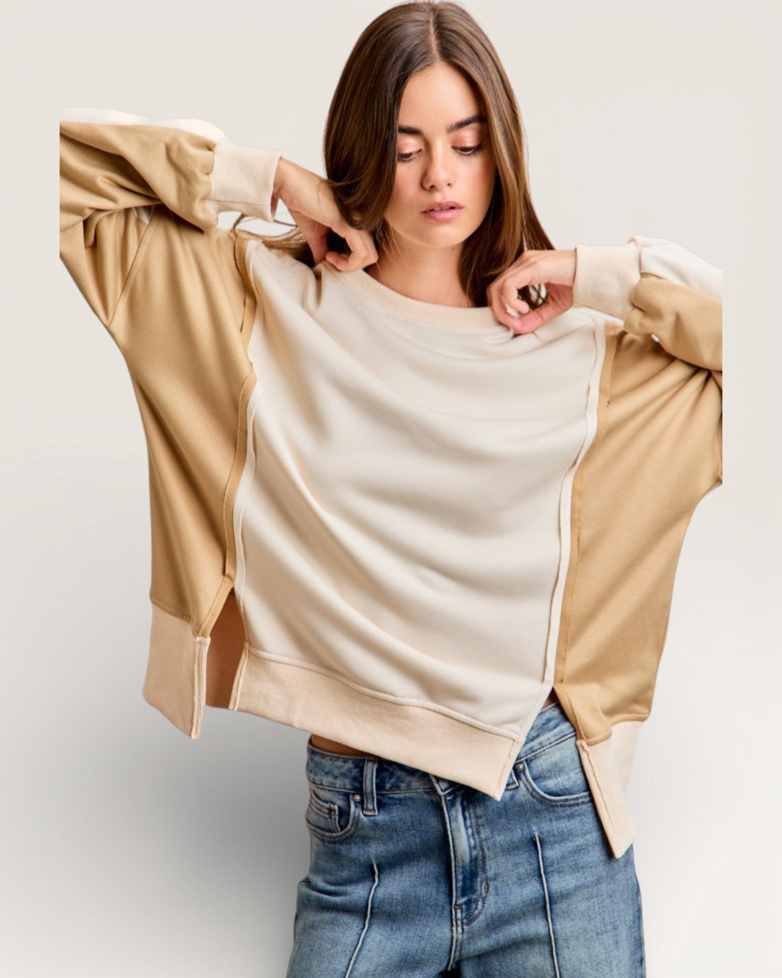 Woman wearing a beige and brown color-blocked sweater with blue jeans on a plain background