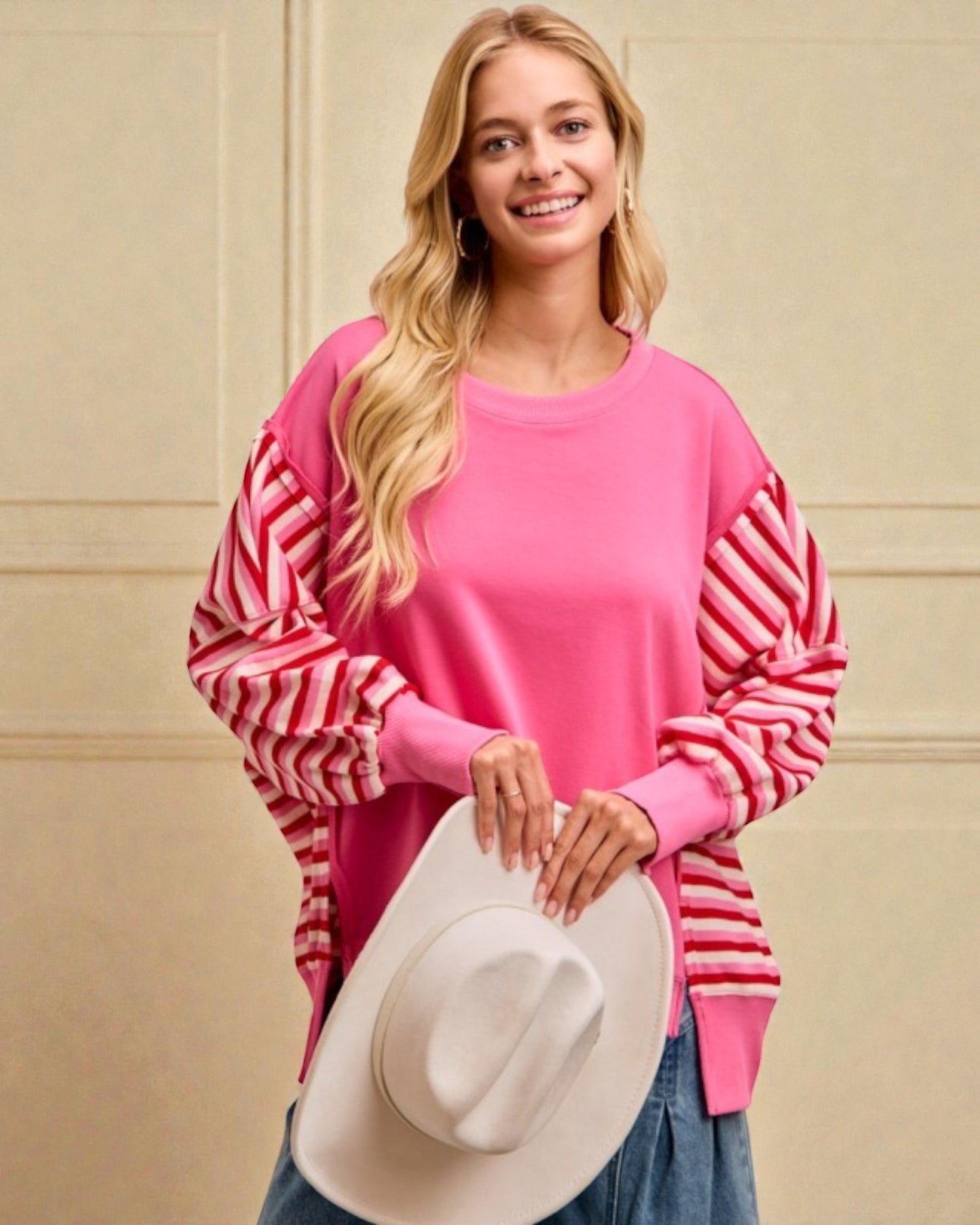 Woman wearing a pink sweater with red stripes, holding a white hat against a beige wall.