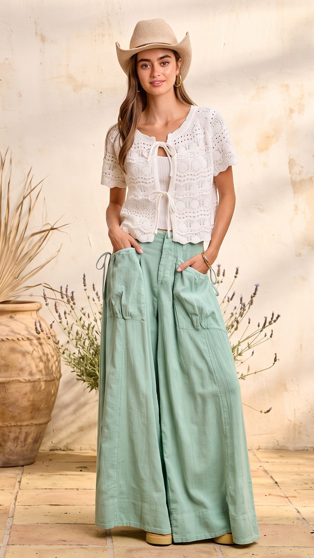 A woman wearing a white crochet short-sleeve top and sage green wide-leg pants stands in a rustic room with a textured beige wall, tile floor, a large clay vase with dried grass, and a small wooden table.