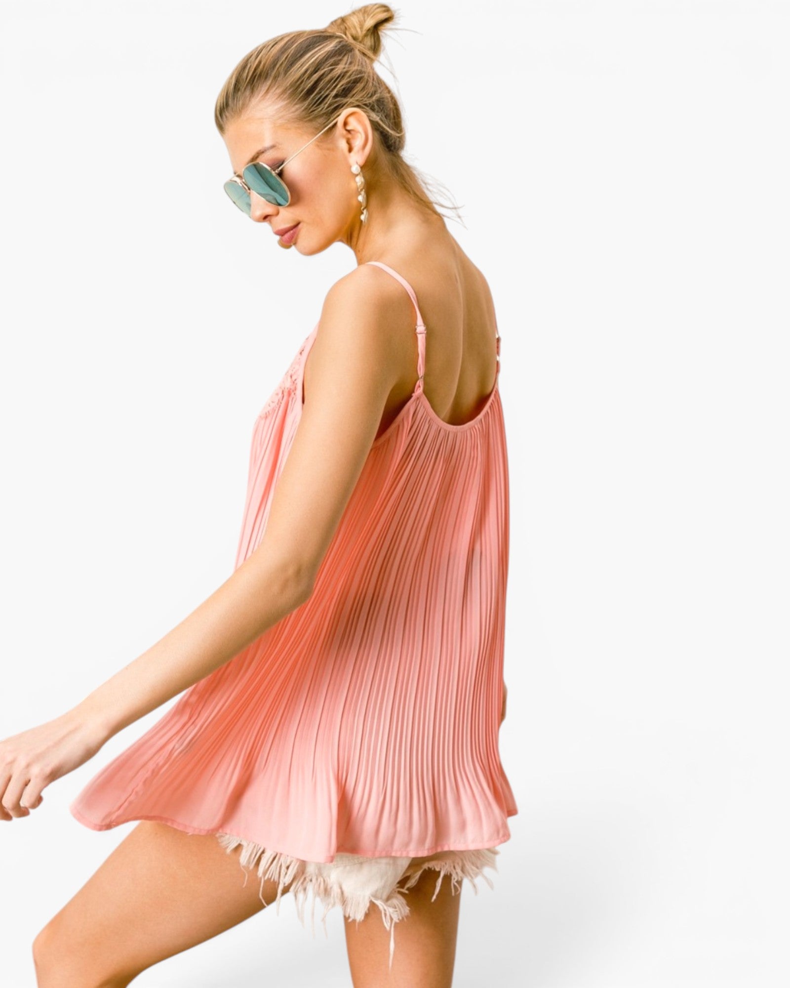 A woman wearing a light pink pleated spaghetti-strap mini dress with a ruffled hem, styled with light blue sunglasses and gold hoop earrings against a plain white background.