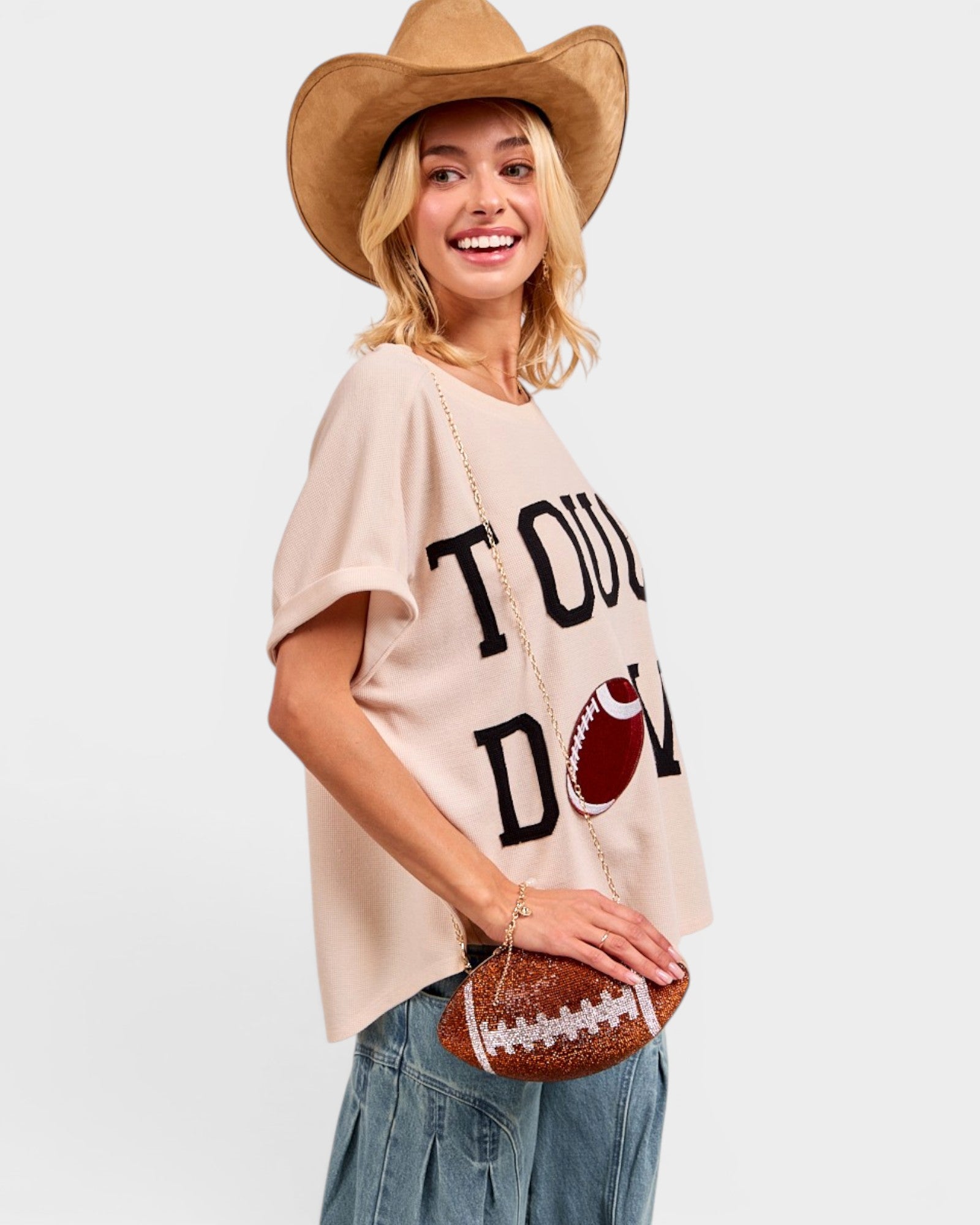 Woman wearing a beige t-shirt with 'Touchdown' text, holding a football, and wearing a cowboy hat on a white background