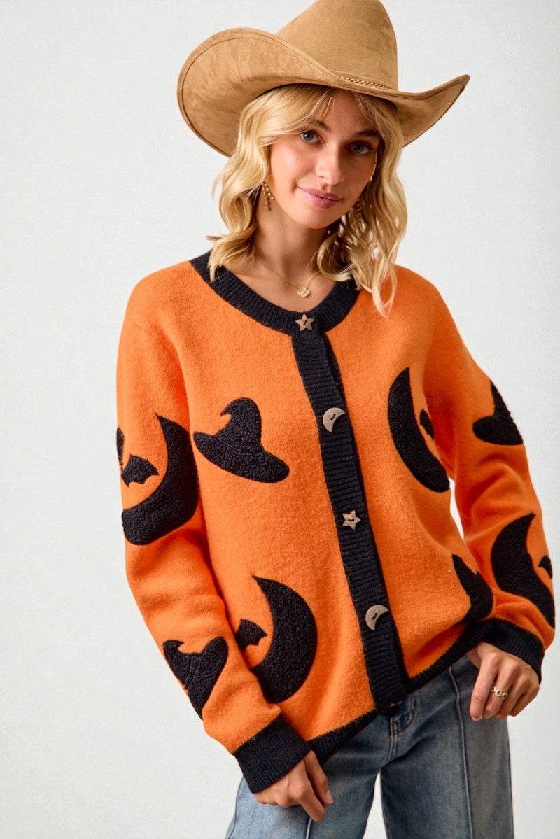 Woman wearing an orange sweater with black witch hat patterns and a cowboy hat.