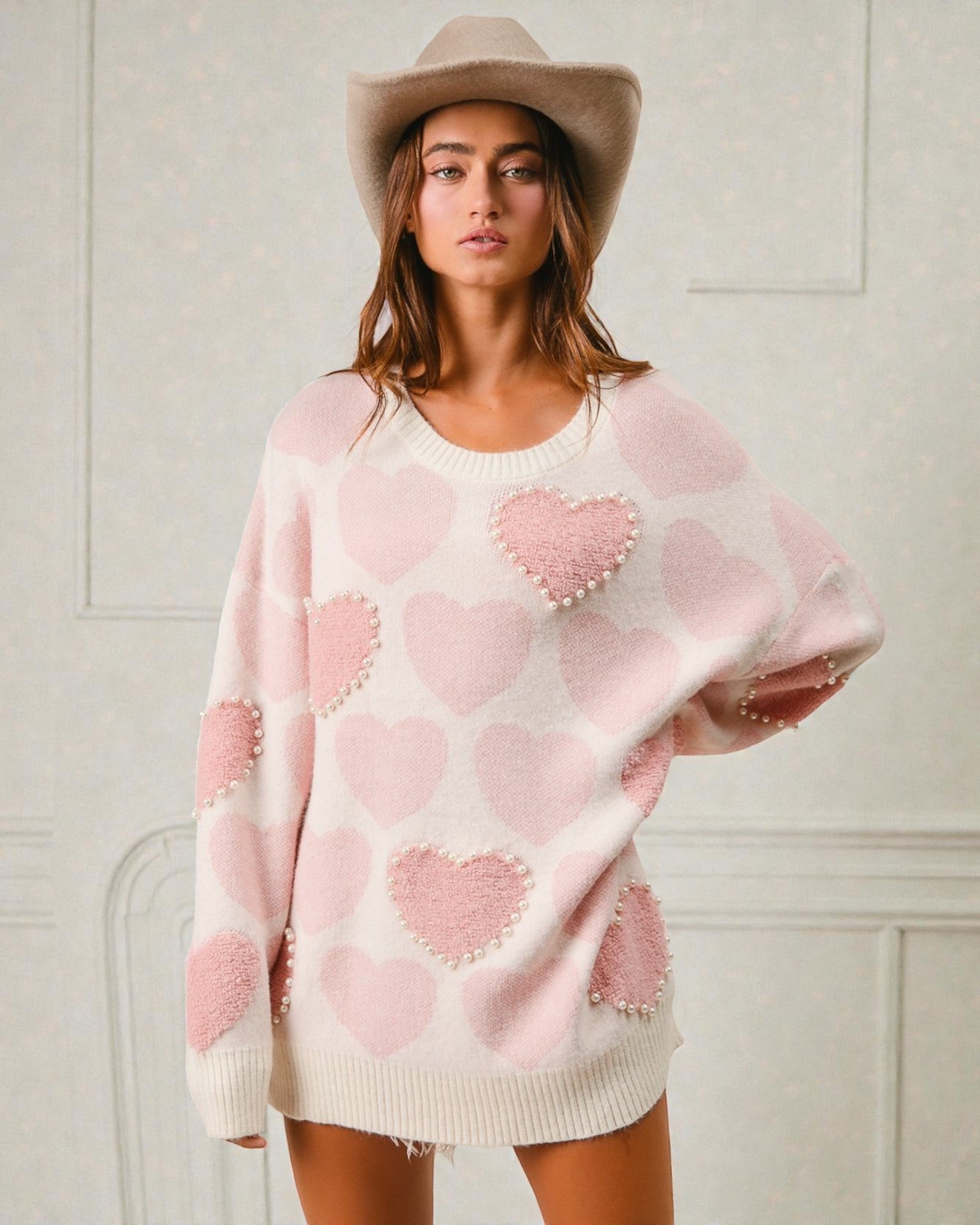 Woman wearing a pink heart-patterned sweater and beige hat against a white wall.