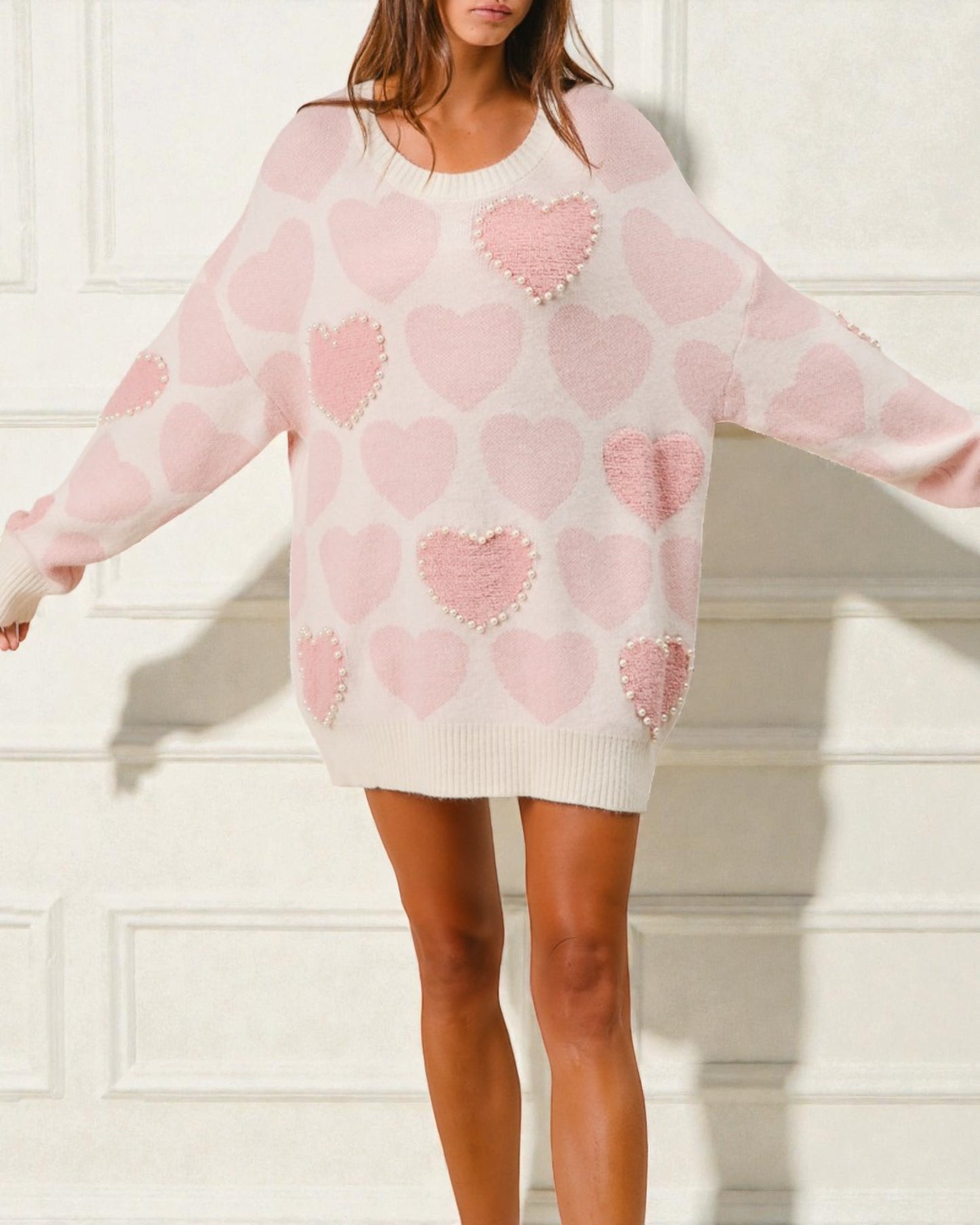 Woman wearing a pink sweater dress with heart patterns against a white wall.