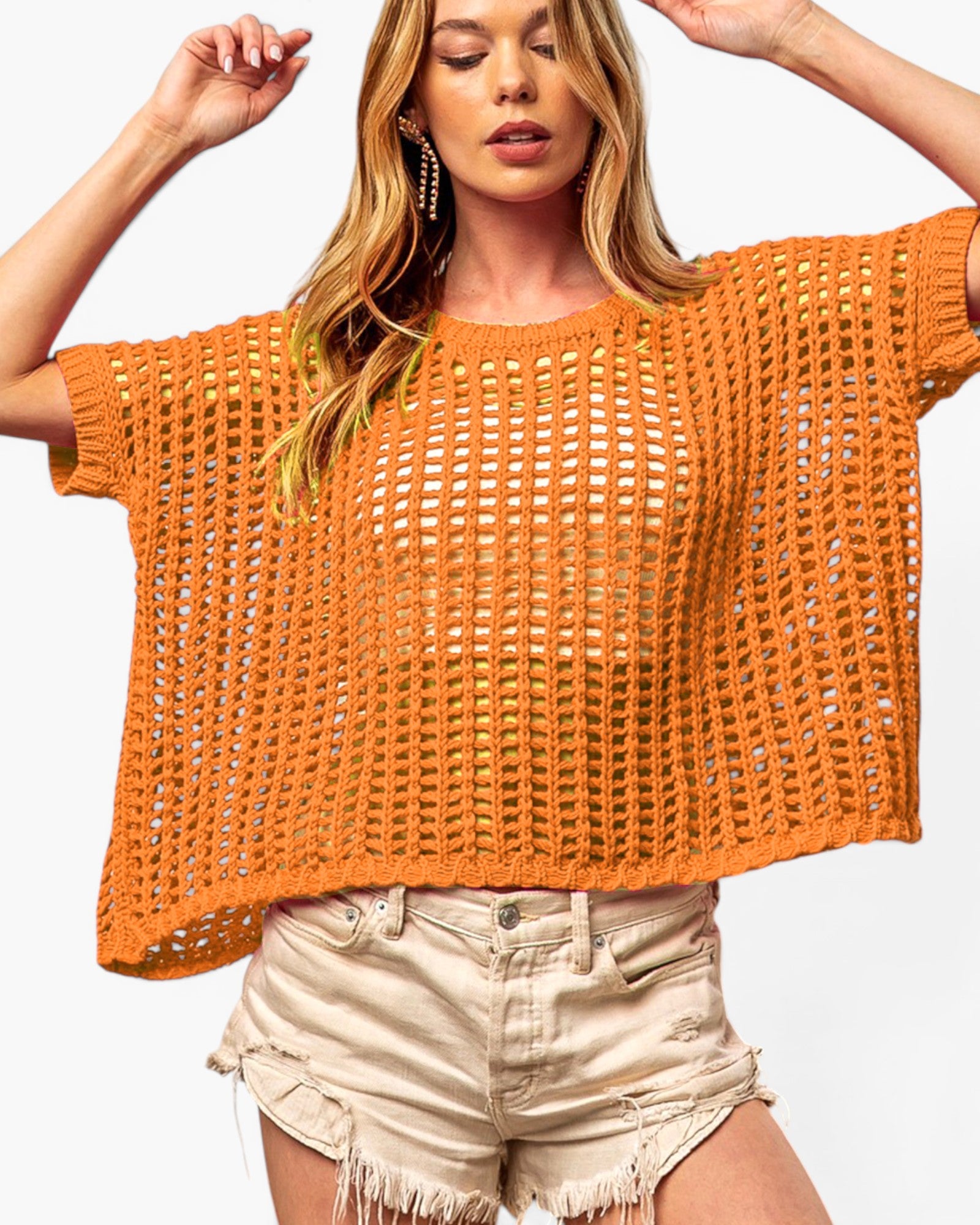 A woman wearing a loose, short-sleeve orange crochet knit top and light beige frayed denim shorts, posing against a plain white background.