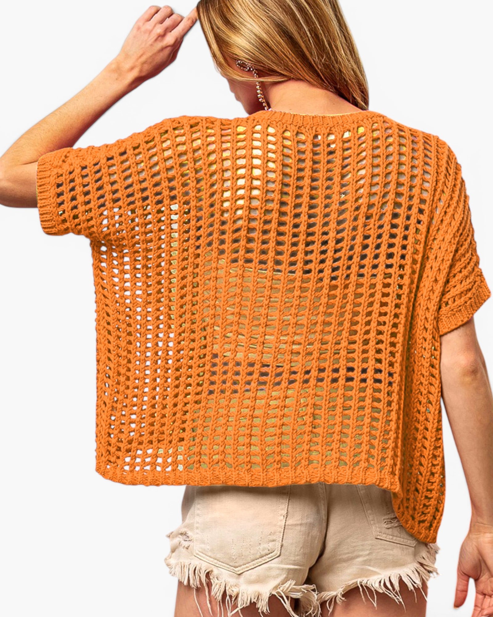 A woman shown from the back wearing a loose, short-sleeve crochet knit top in a warm orange-brown color, paired with light beige frayed denim shorts against a plain white background.