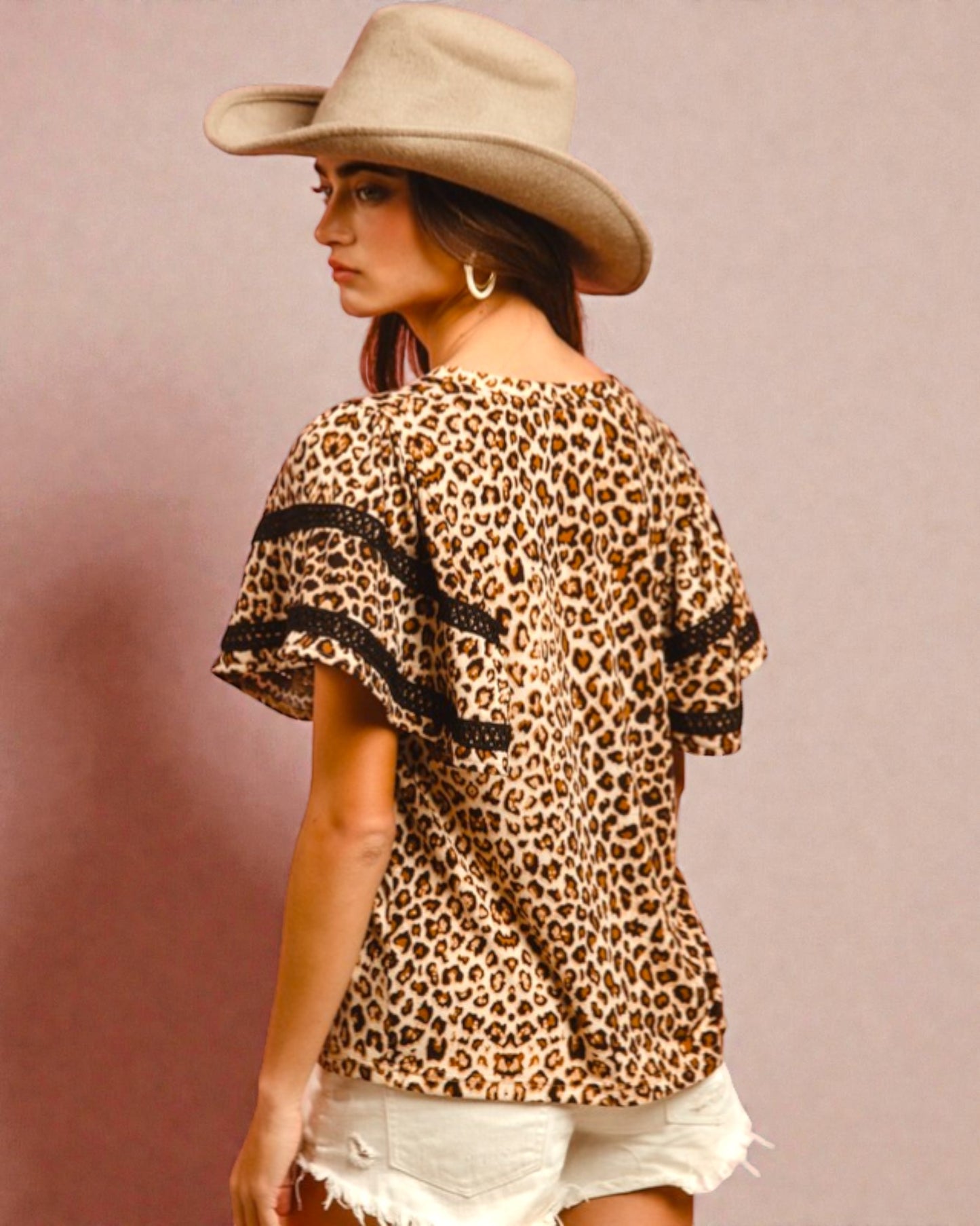 Woman wearing a leopard print top with black trim and a wide-brimmed hat on a pink background