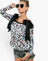 A woman wearing a long-sleeve leopard print top with black bow details at the shoulders, paired with denim shorts and sunglasses, against a plain light gray background.