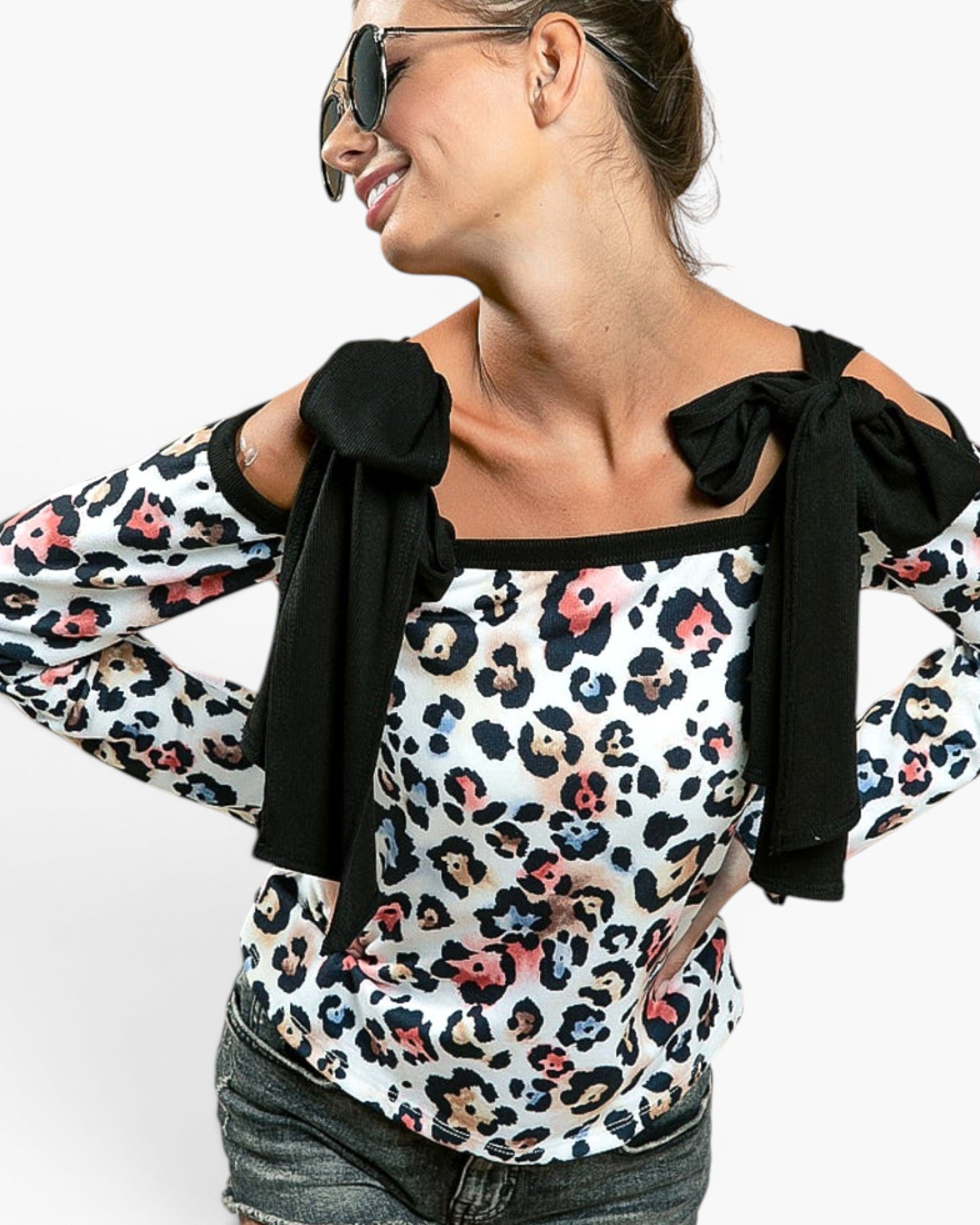 A woman wearing a white long-sleeve top with a multicolor leopard print and black bow details on the shoulders, paired with dark gray jeans and sunglasses, against a plain white background.