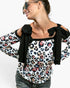 A woman wearing a white long-sleeve top with a multicolor leopard print and black bow details on the shoulders, paired with dark gray jeans and sunglasses, against a plain white background.