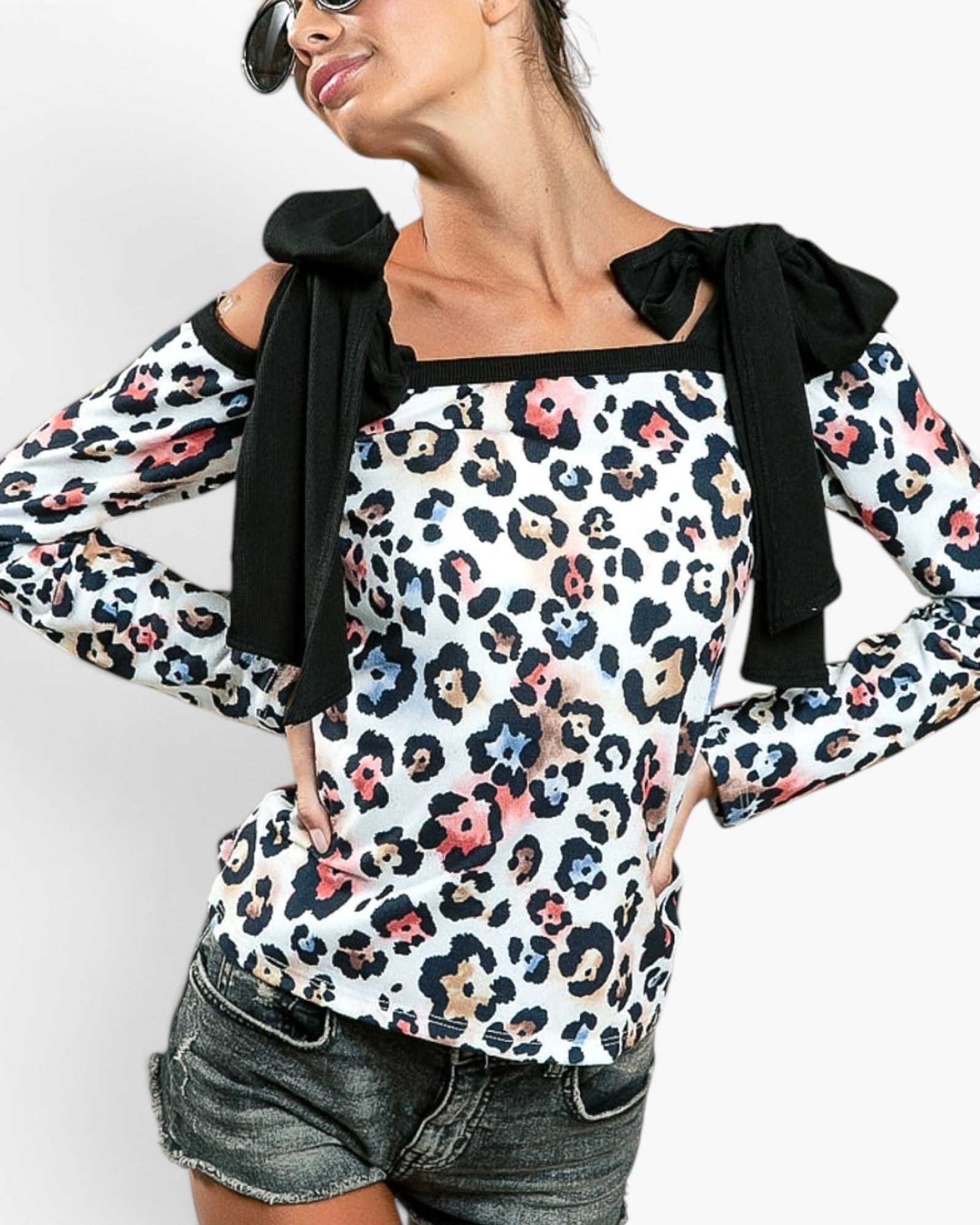 A woman wearing a long-sleeve leopard print top with black bow details at the square neckline and black trim, paired with denim shorts, against a light gray studio background.
