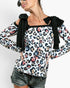 A woman wearing a long-sleeve leopard print top with black bow details at the square neckline and black trim, paired with denim shorts, against a light gray studio background.