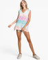 A woman modeling a sleeveless V-neck tunic top with a pastel leopard print pattern in pink, light blue, and white, paired with denim shorts and white sneakers against a light gray studio background.