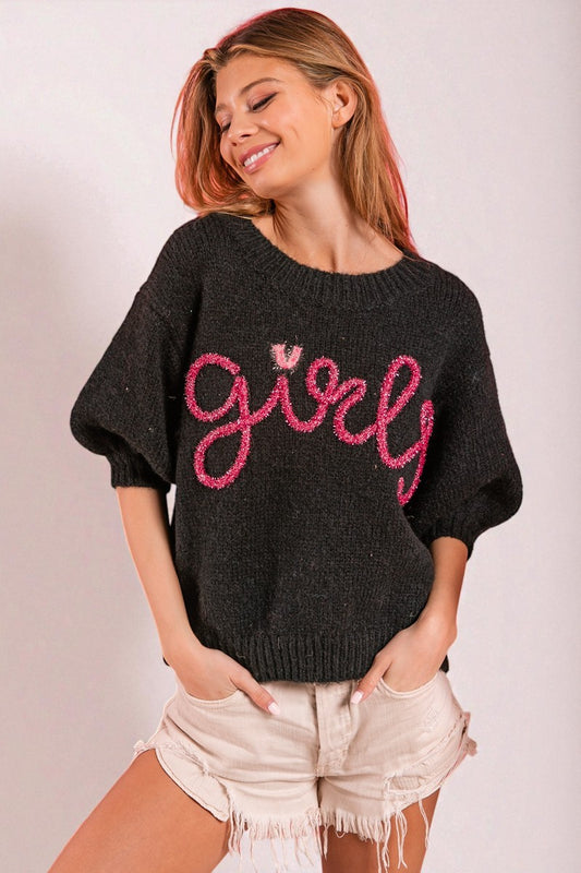 Woman wearing a black sweater with pink text and white shorts on a light background