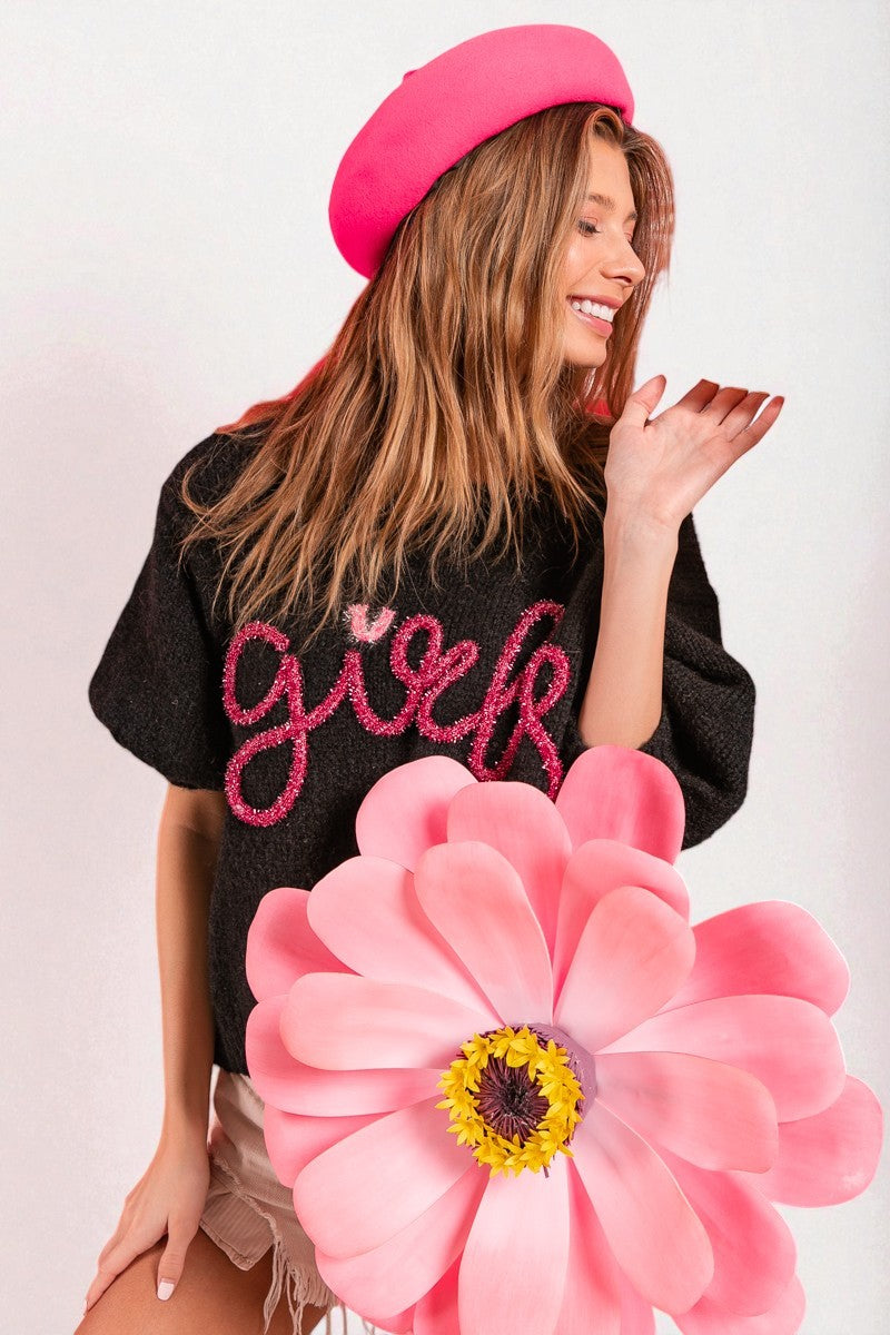 Woman wearing a black sweater with pink text, pink beret, and holding a large pink flower against a white background