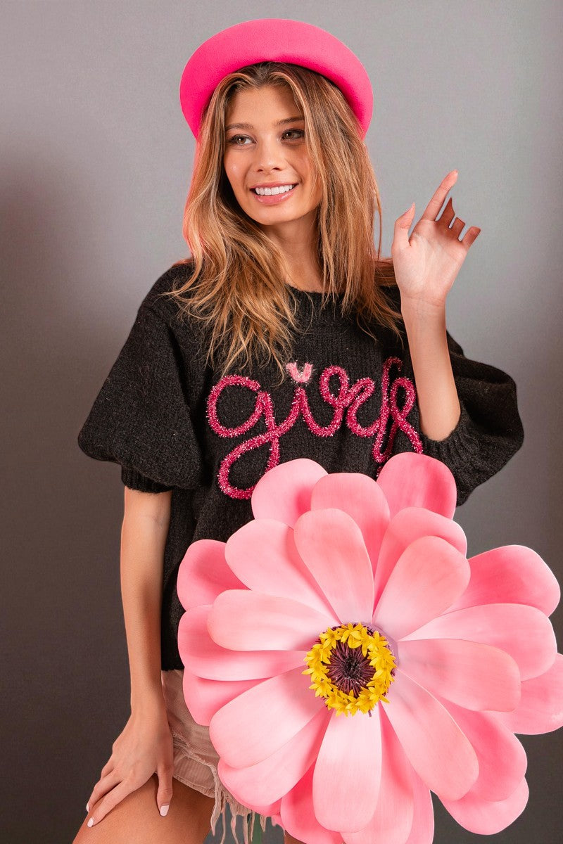 Woman wearing a black t-shirt with 'girly' text, pink beret, and large pink flower in front of a gray background