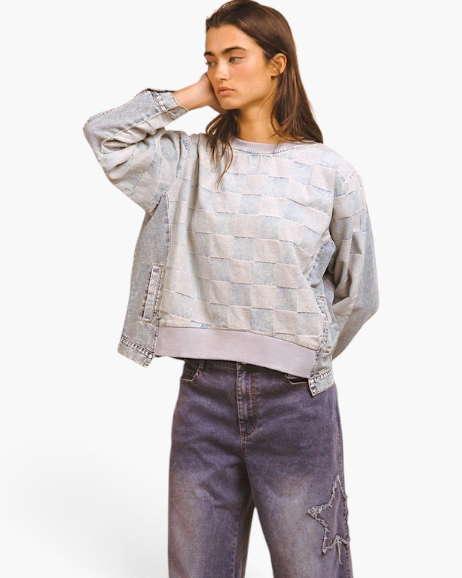 A woman modeling a cropped, light-wash denim jacket with a checkerboard patchwork pattern and ribbed hem, paired with dark denim jeans, against a plain white studio background.